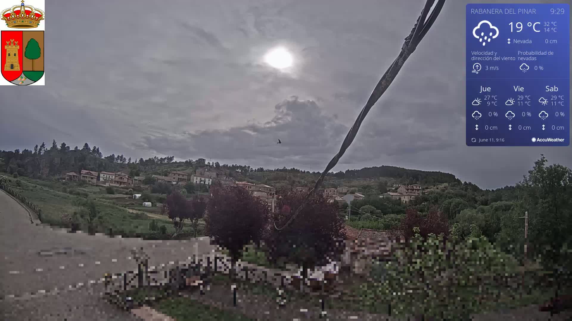 A small village nestled on a hillside is viewed under an overcast sky with a bright sun partially obscured by clouds, a bird flying overhead.