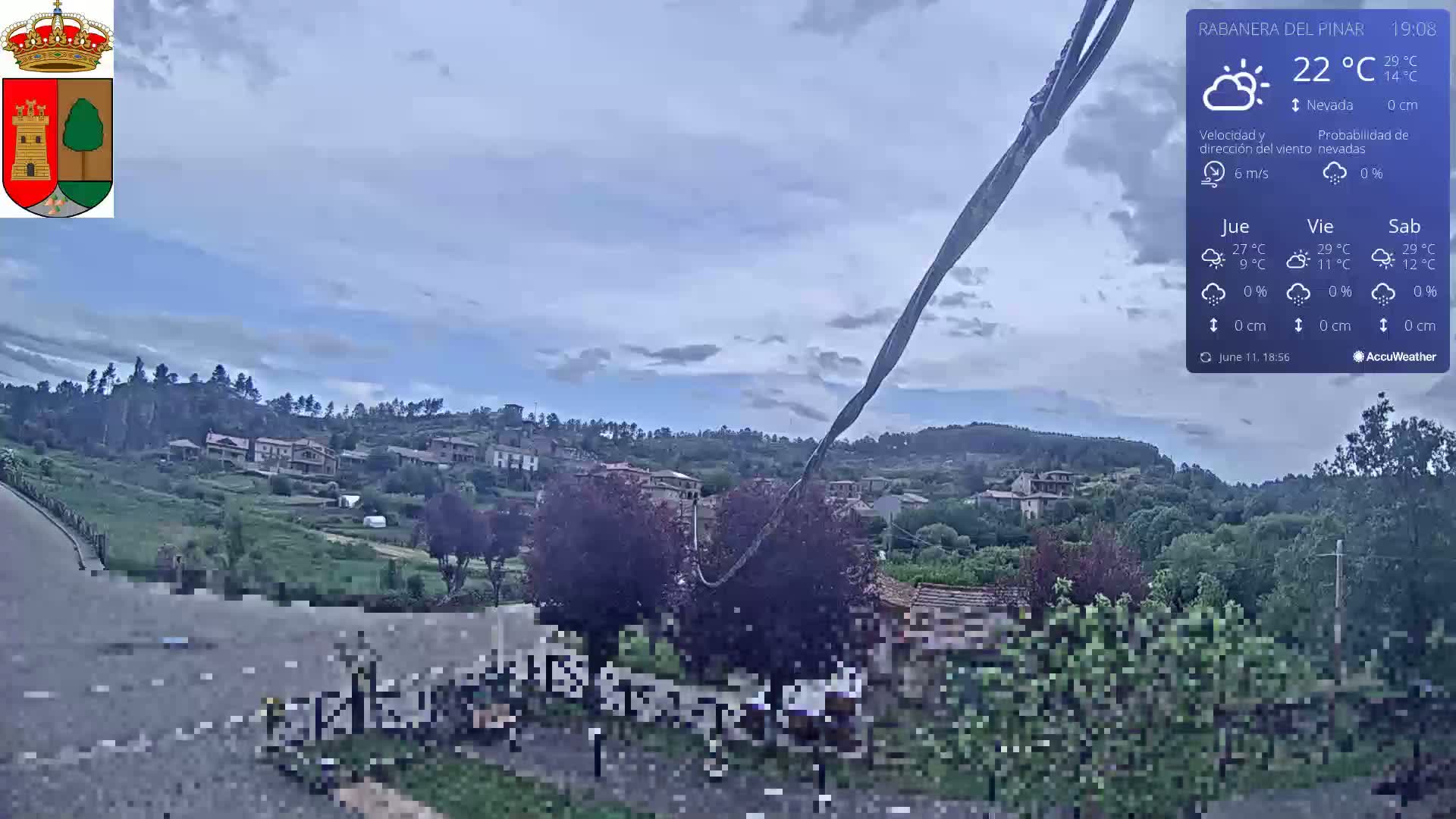 A mostly cloudy view of a small village nestled in a hilly, green landscape under a partly cloudy sky.