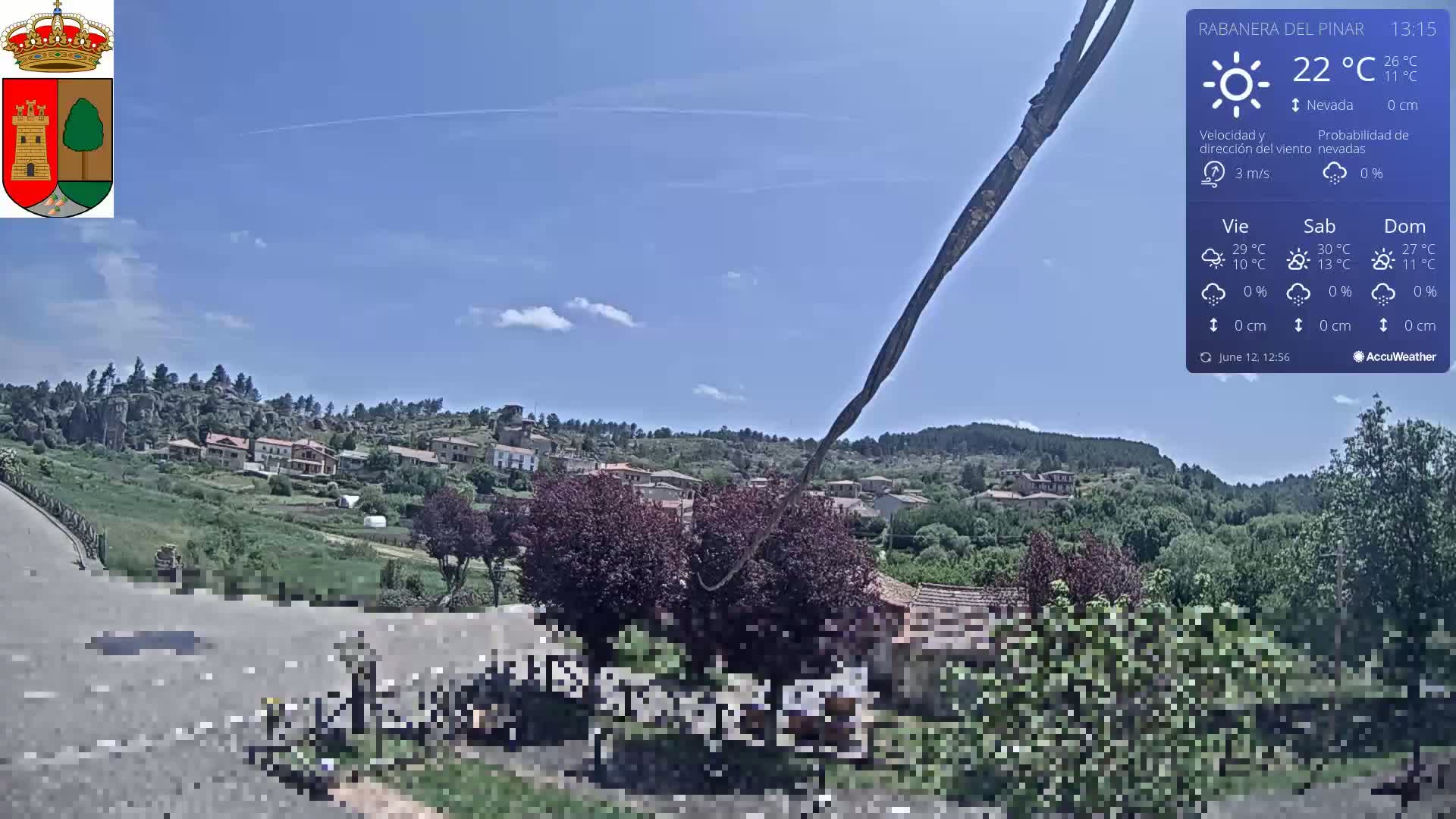A sunny, partly cloudy view of a small village nestled in a hilly, green landscape.