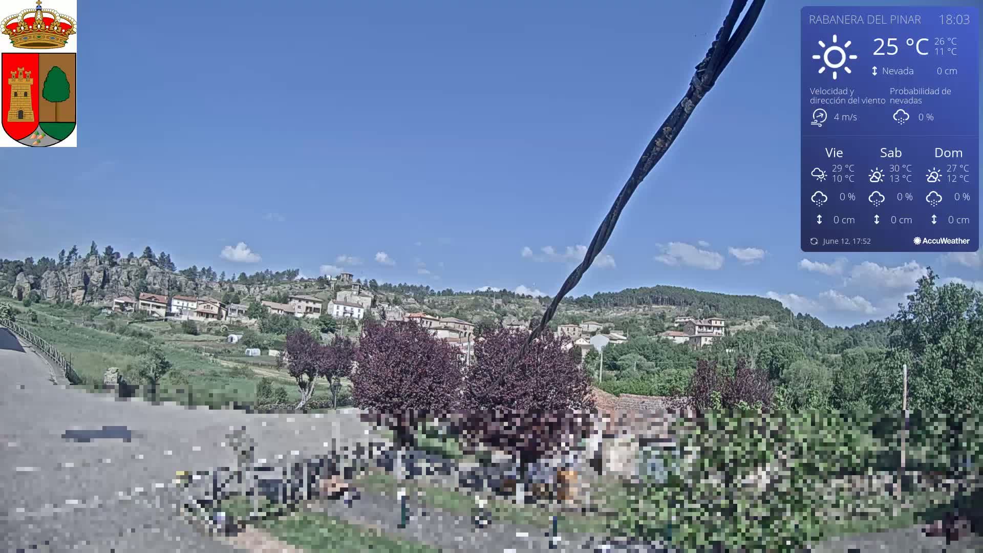 A mostly sunny view of a small village nestled among hills and trees, under a clear blue sky.