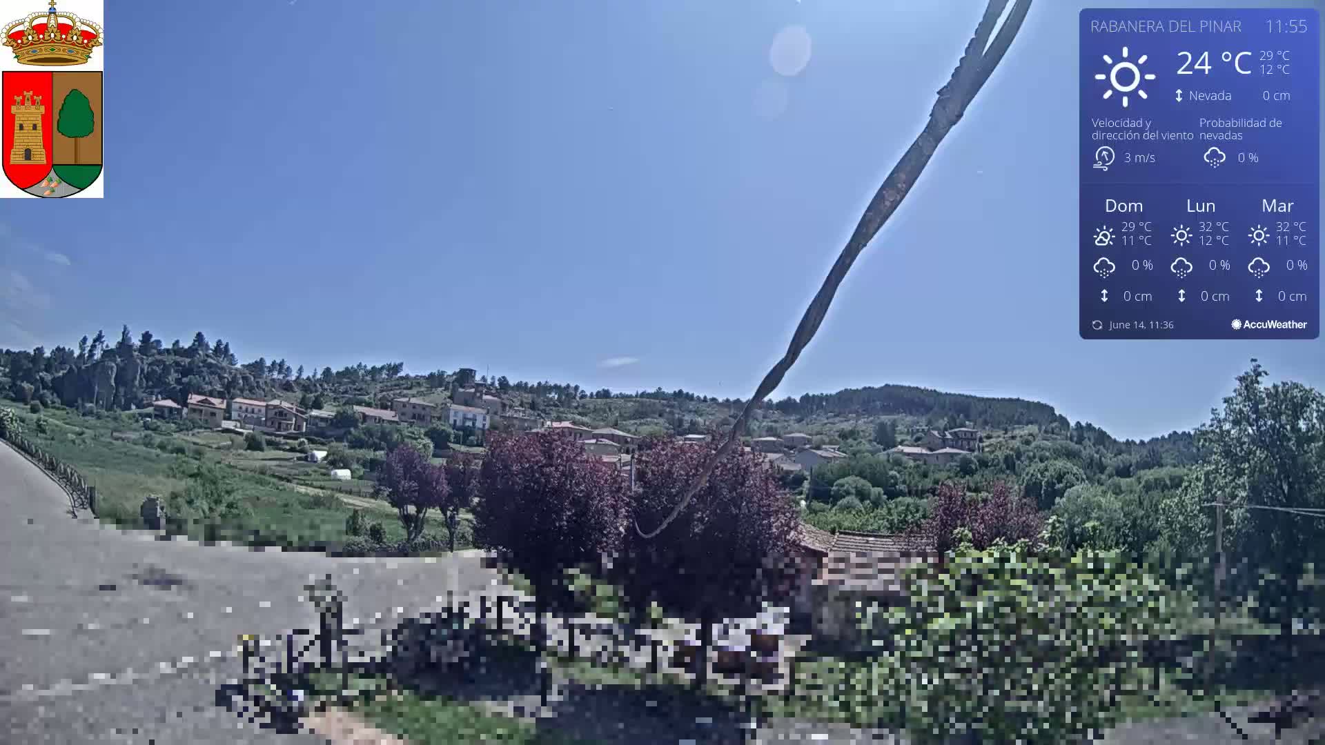 A sunny view of a small village nestled among green hills and trees, under a clear blue sky.
