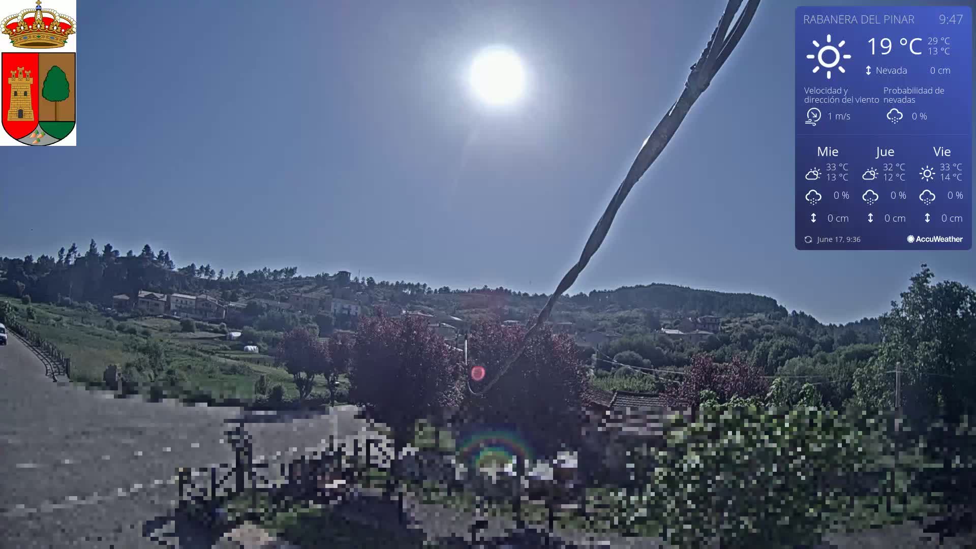 A sunny, clear day overlooks a small village nestled in a hillside, with greenery and a road visible in the foreground.