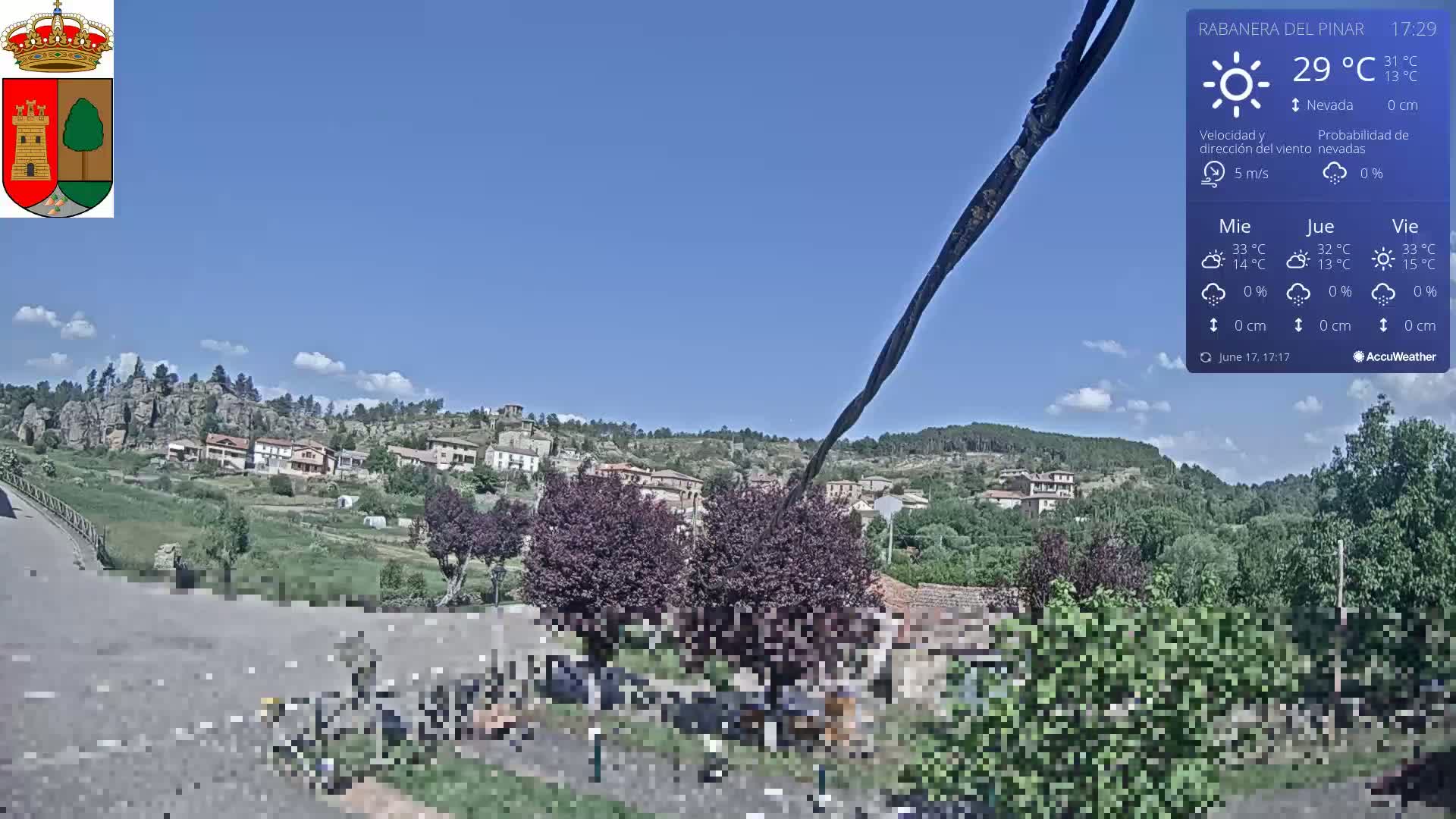 A small village nestled on a hillside under a mostly clear, bright blue sky is viewed from a slightly elevated position, with some scattered clouds visible.
