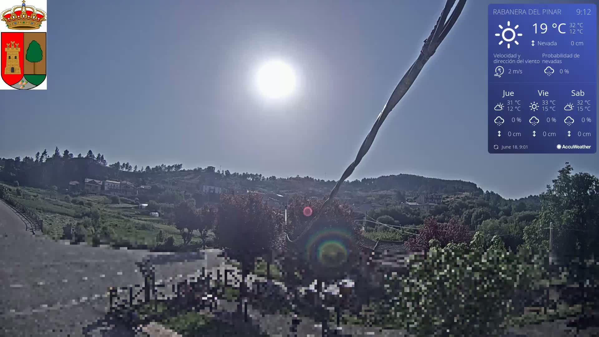 A sunny, clear day shows a small village nestled in a hillside valley, with a bright sun overhead and some vegetation in the foreground.