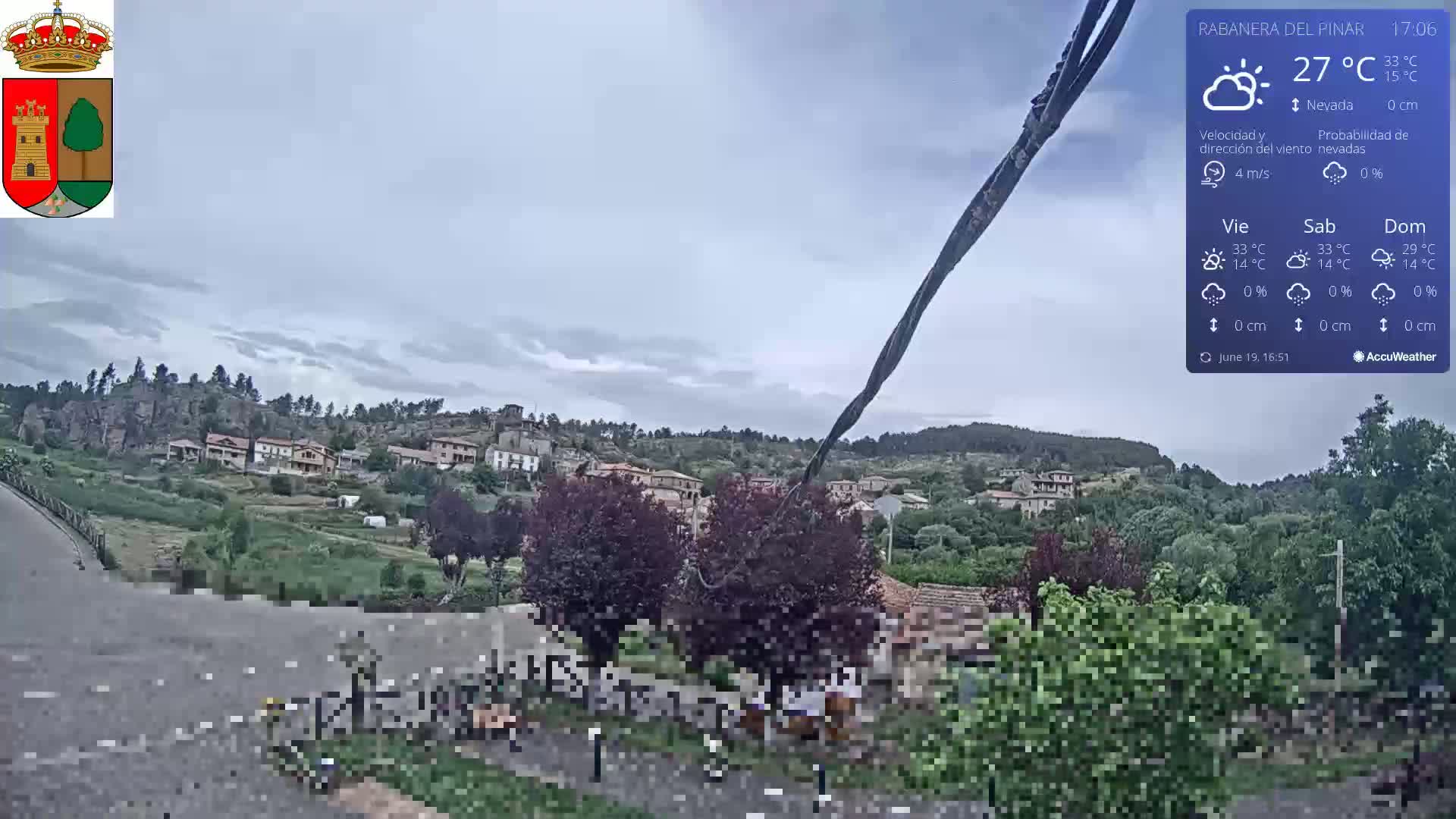 A partly cloudy day reveals a small village nestled on a hillside, with vegetation and a road in the foreground.