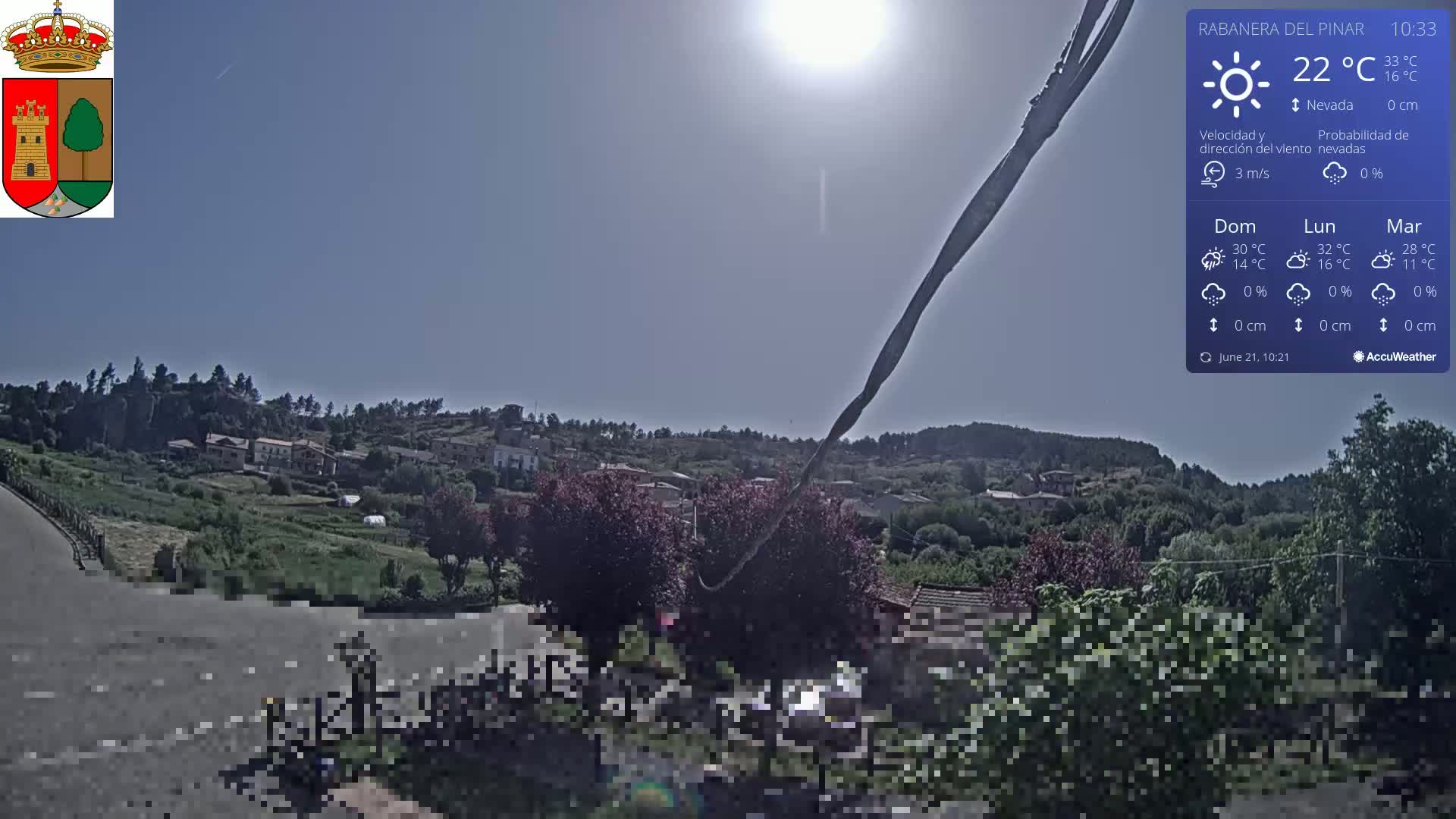 A sunny day reveals a small village nestled among green hills, with a clear blue sky and a few trees in the foreground.
