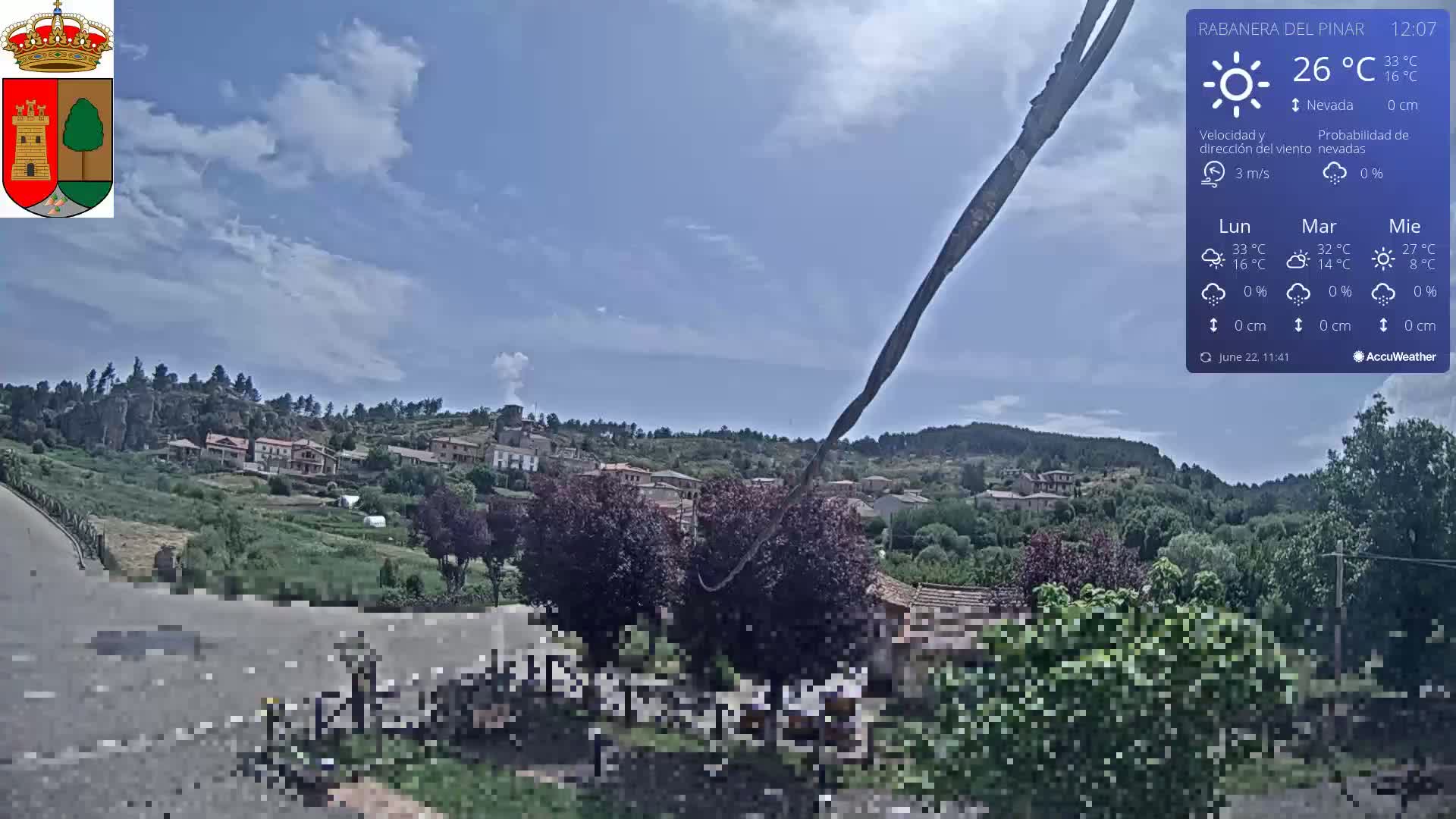 A partly cloudy day reveals a small village nestled in a green hillside, viewed from a slightly elevated position along a road.