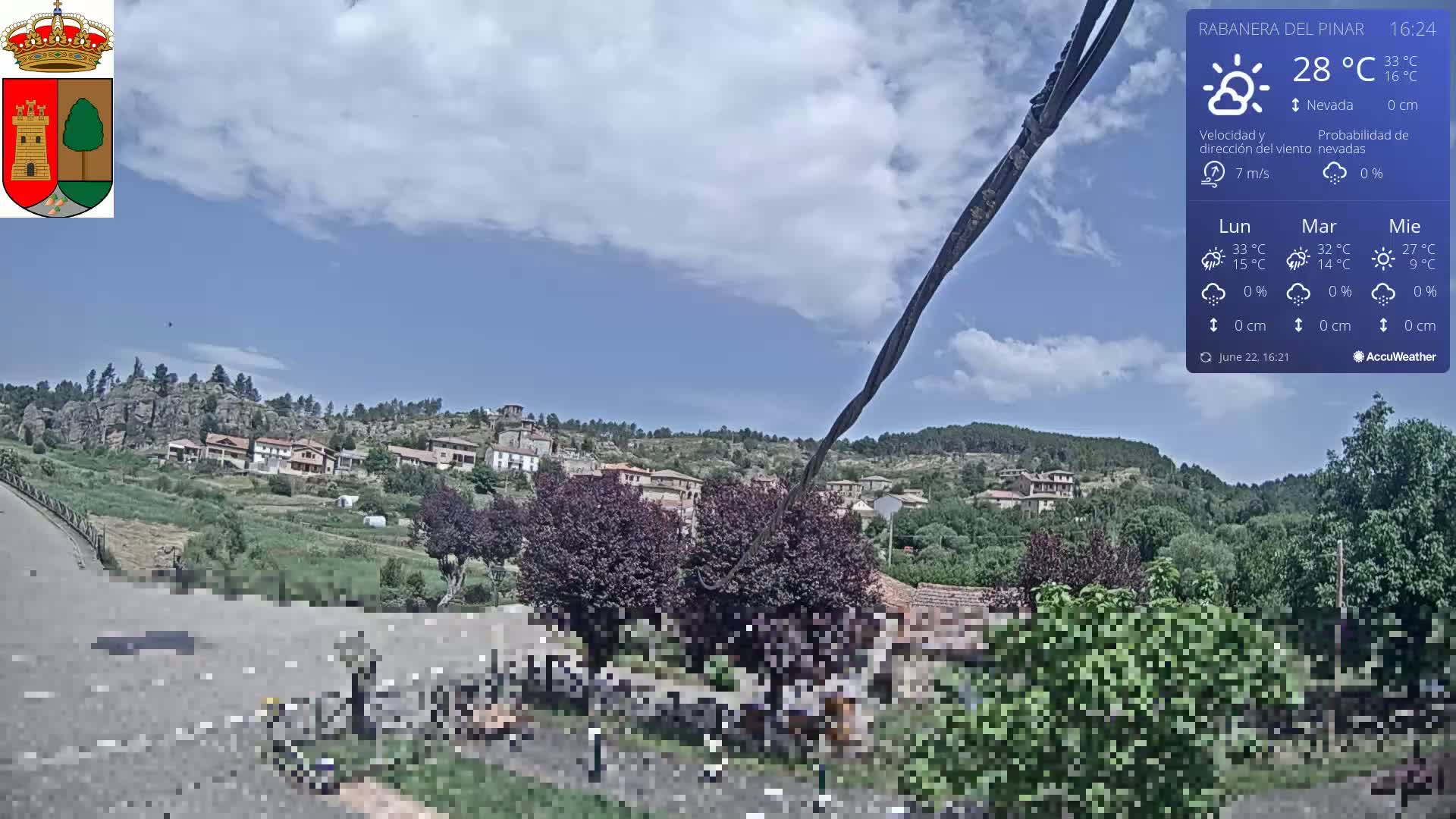 A partly cloudy sky over a small village nestled in a hillside, with green vegetation and rocky outcrops.