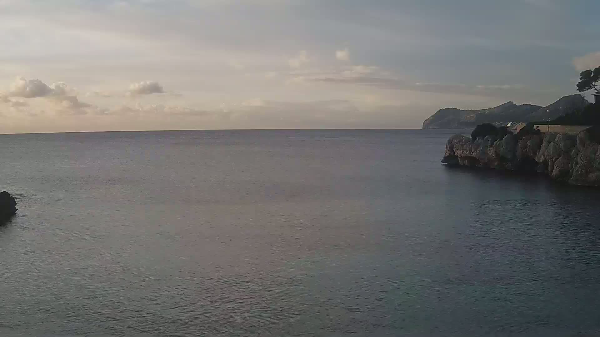 A calm, dark-blue sea stretches to a distant, hazy coastline under a mostly cloudy, pale sky.