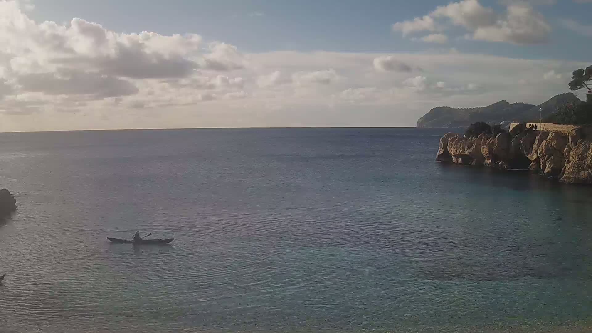 A person kayaks in calm, clear water near a rocky coastline under a partly cloudy sky.