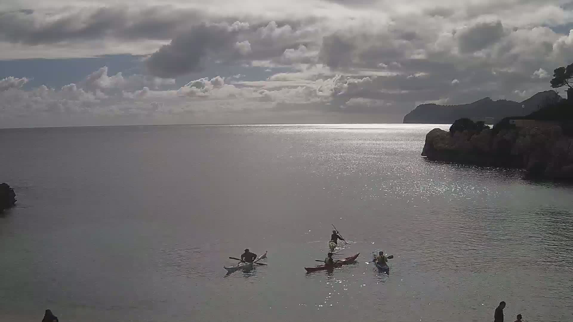 Several people kayak on a calm sea under a cloudy sky near a rocky shore and distant mountains.