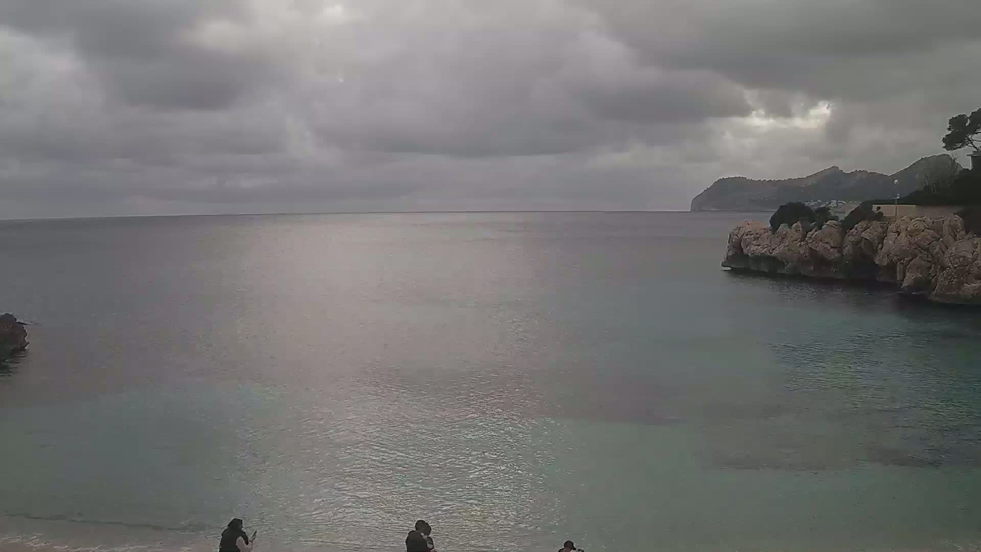A calm, grey-toned sea laps a sandy shore under a cloudy sky, with a few people visible near the water's edge and rocky cliffs in the background.