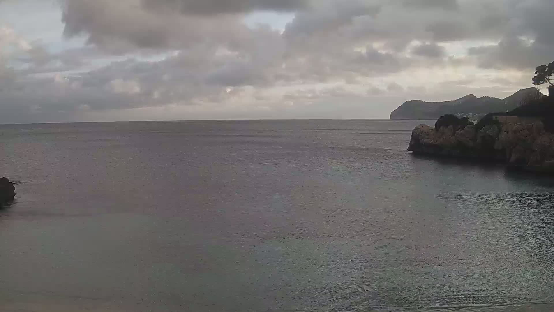 A calm, gray sea laps against rocky shores under a cloudy sky, with a distant landmass visible in the background.