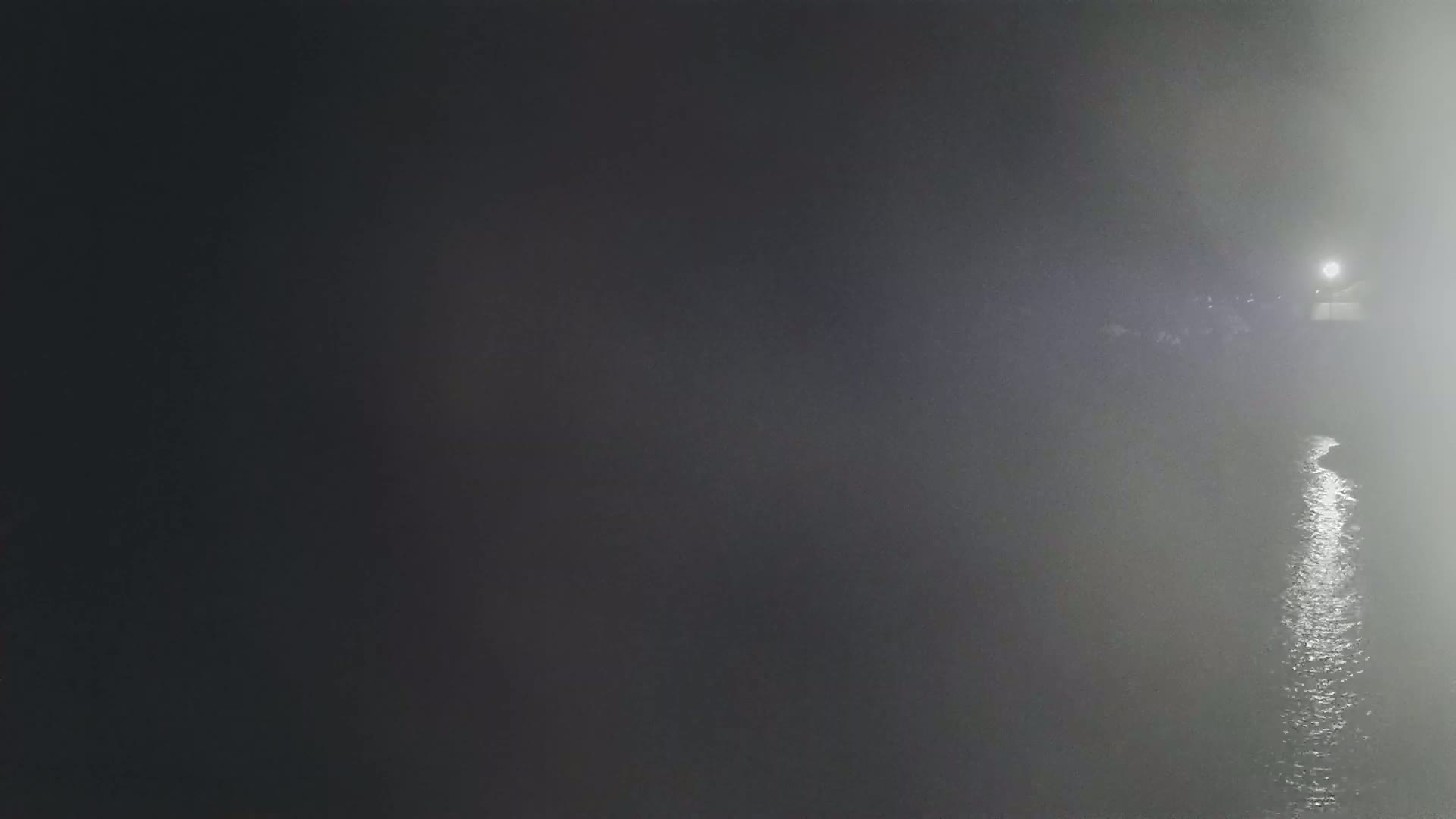 A foggy night scene shows a body of water reflecting light, with indistinct shapes and a light source visible in the distance.