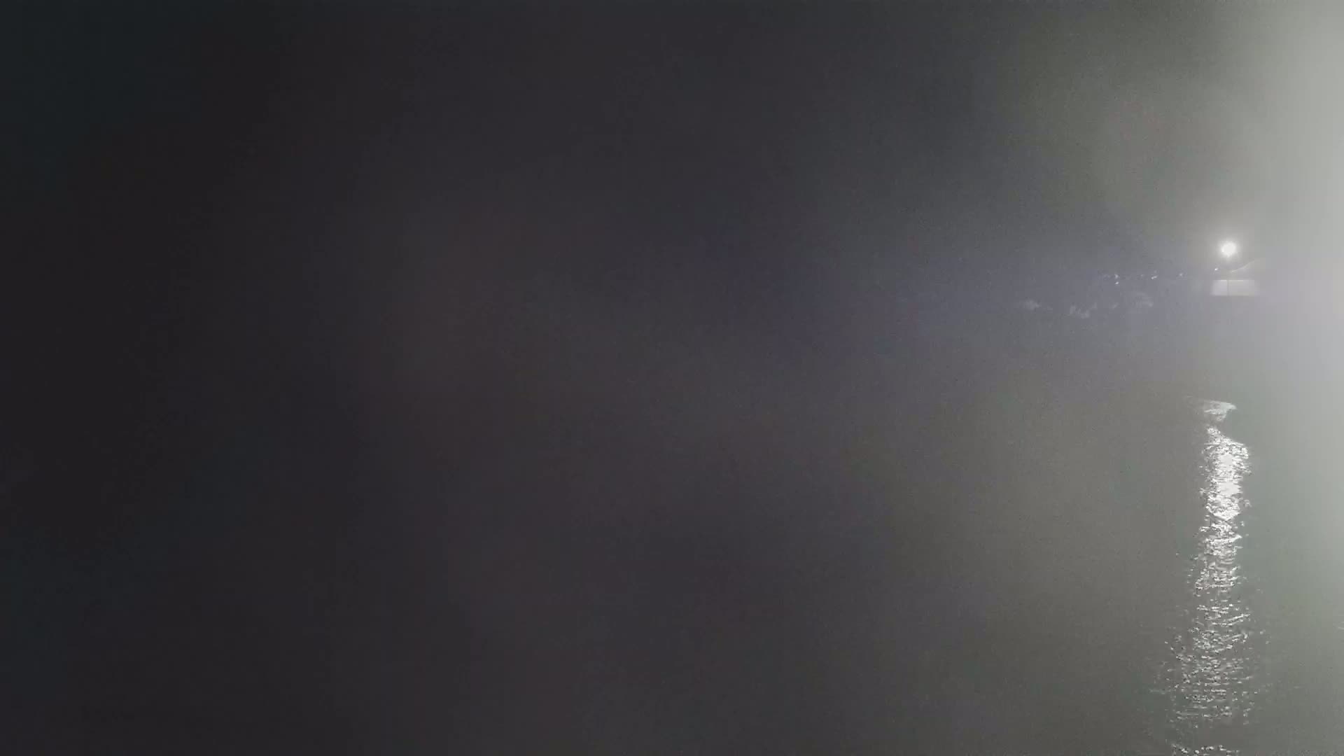 A body of water is visible at night in foggy conditions, with a light source reflecting on its surface in the distance.