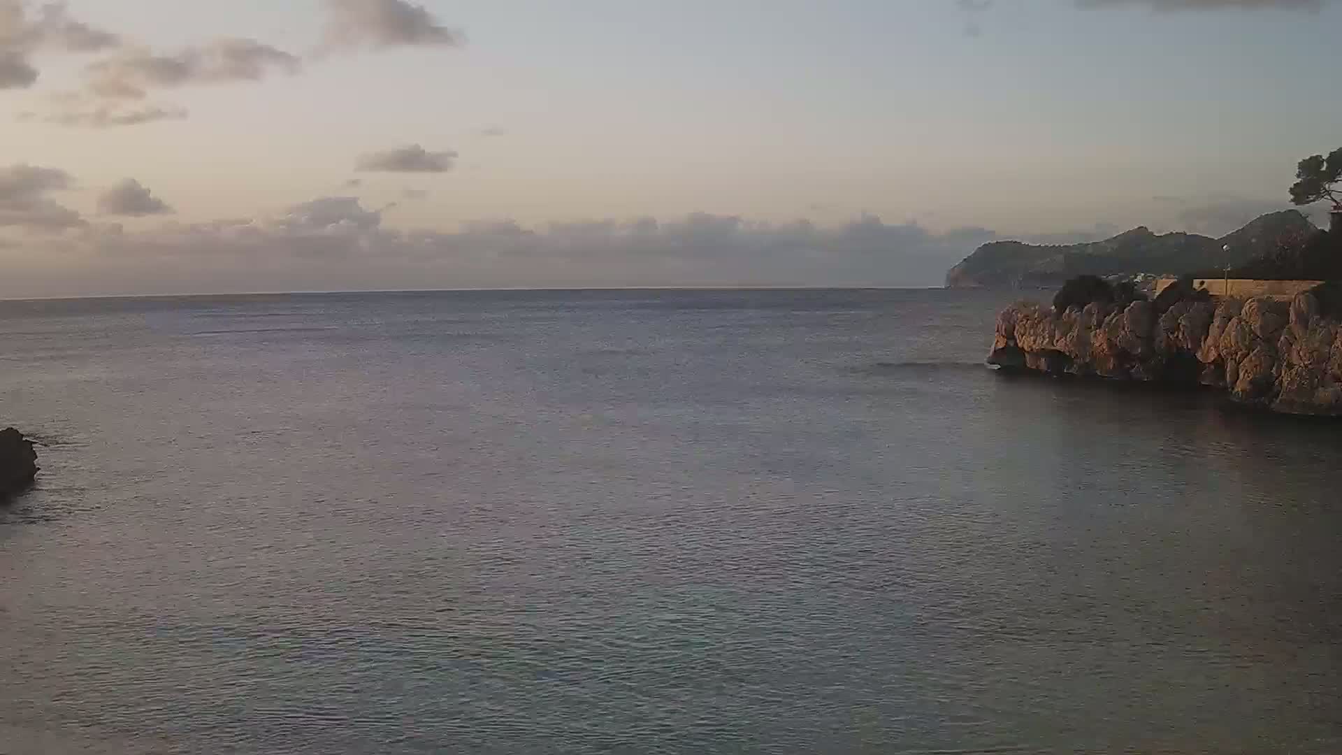 A calm sea under a cloudy sky is partially enclosed by rocky cliffs, with land visible in the distance.