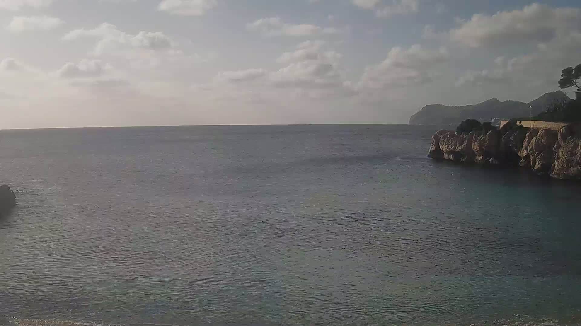 A calm sea under a partly cloudy sky is seen from a rocky cove, with a distant mountain visible on the horizon.