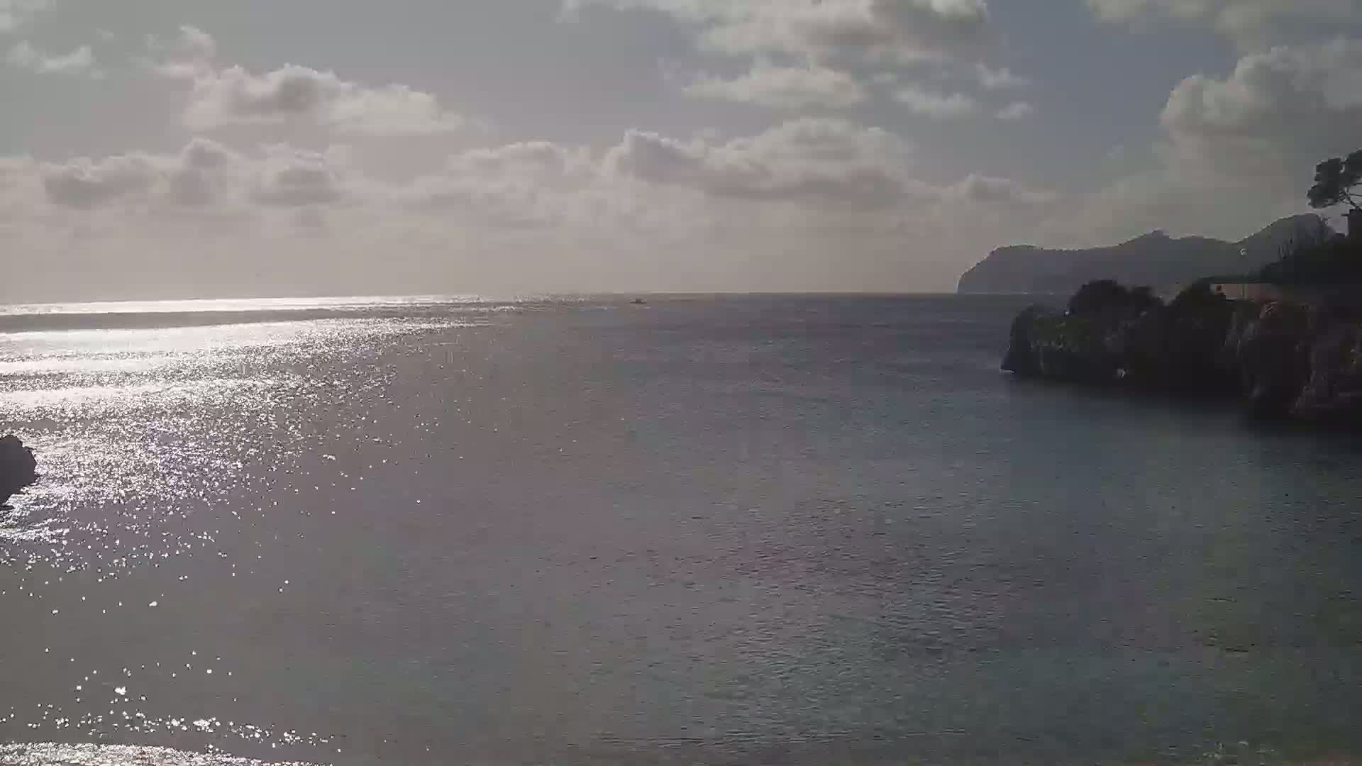 A calm, sparkling sea under a partly cloudy sky is seen from a coastal vantage point, with a distant landmass visible in the background.