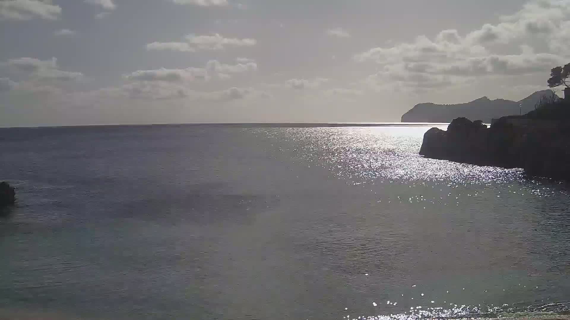 A calm body of water shimmers under a partly cloudy sky, with dark rocky shorelines and a distant mountain visible.