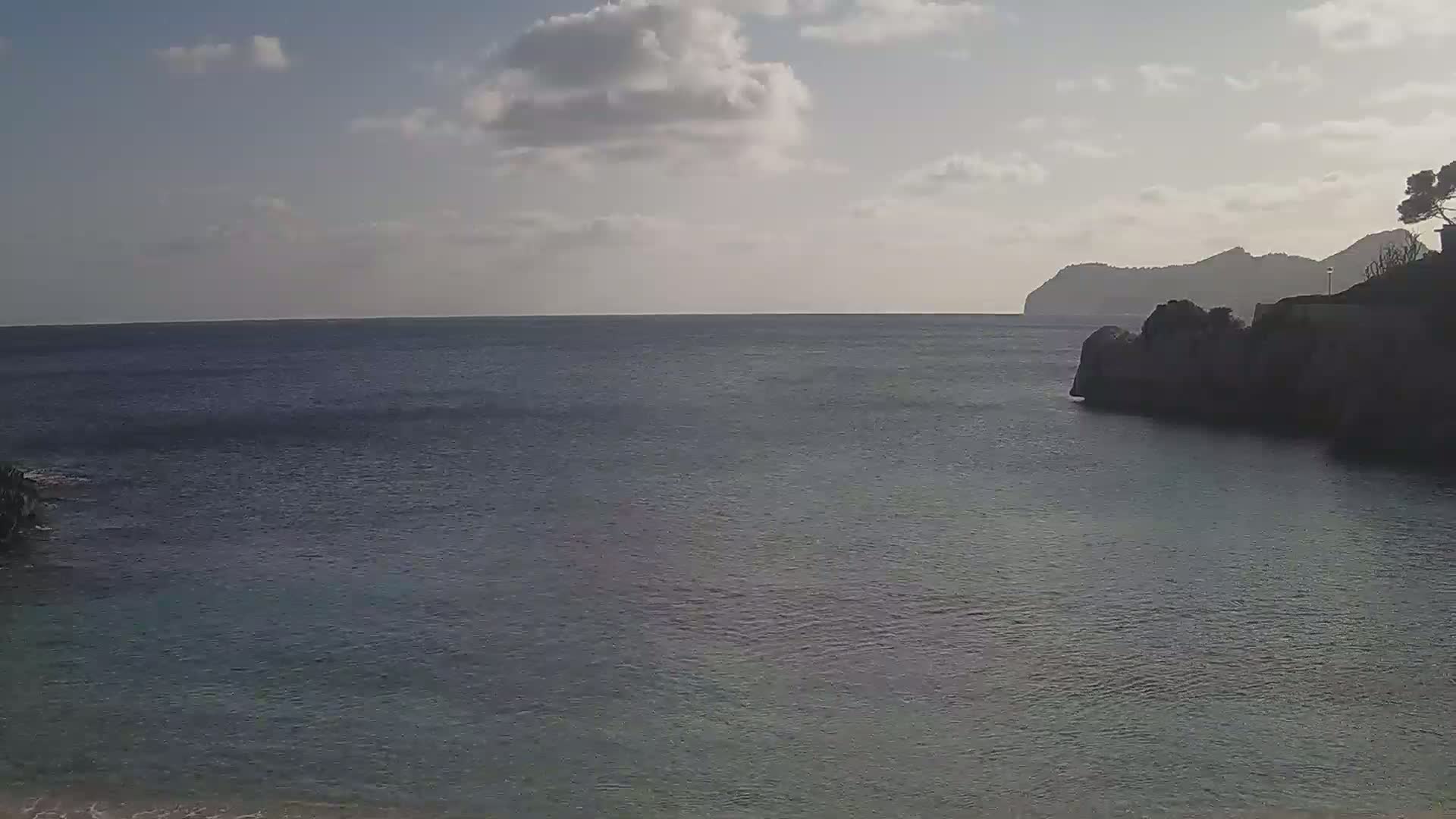 A calm, partly cloudy day shows a dark-colored body of water, partially enclosed by rocky land with sparse vegetation.