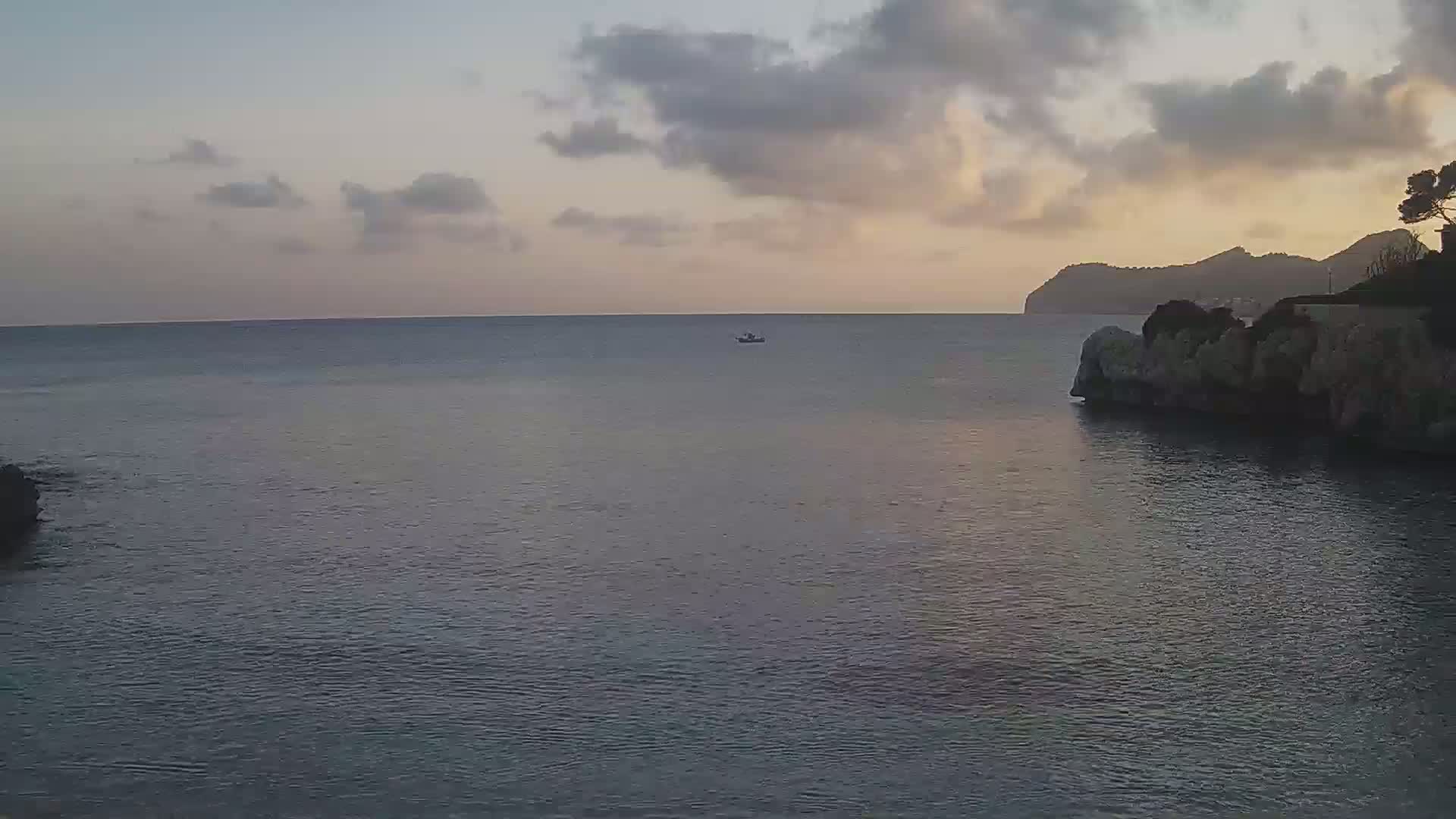 A calm sea at sunset, under a partly cloudy sky, with a small boat in the distance and rocky shores.