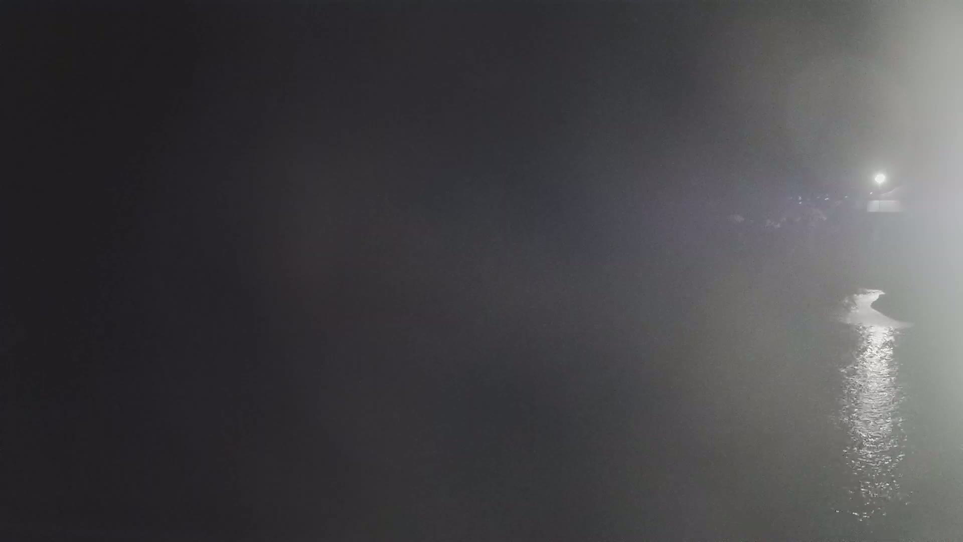 A dimly lit, foggy scene shows a body of water reflecting light, with a group of indistinct figures visible in the distance on the right.