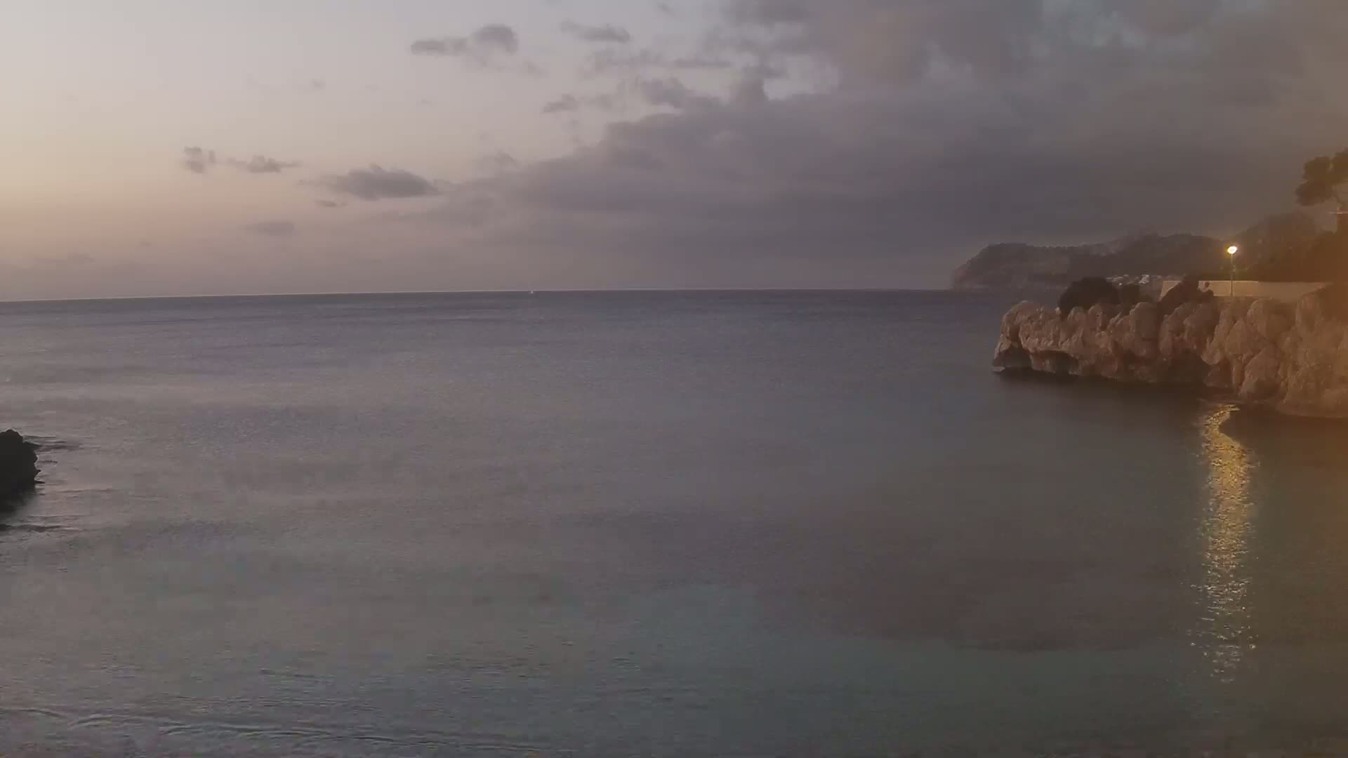 A calm sea at dusk, with a rocky shoreline and a distant hill under a cloudy sky.