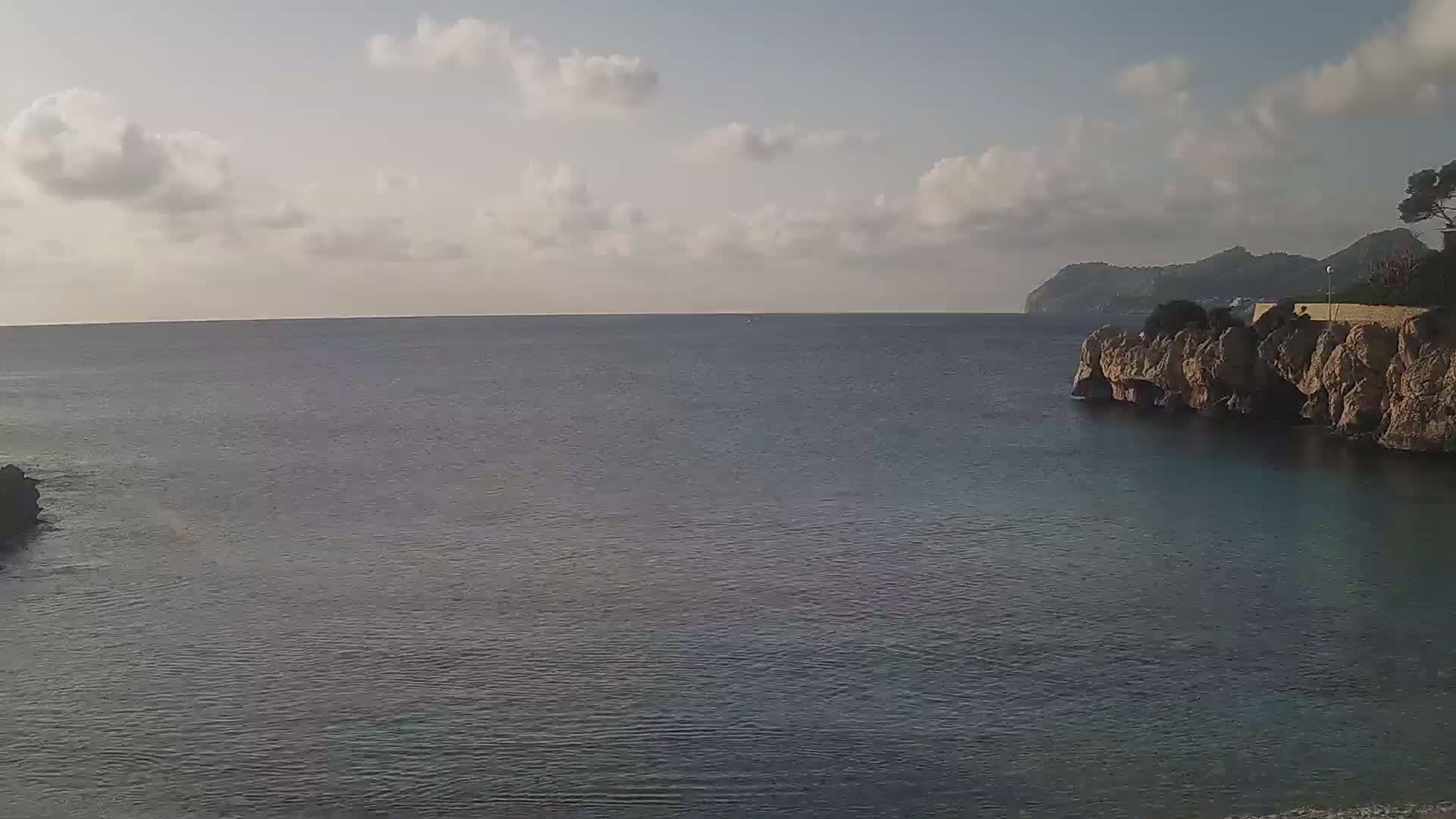 A calm, partly cloudy day shows a rocky coastline with a calm sea stretching to a distant landmass.