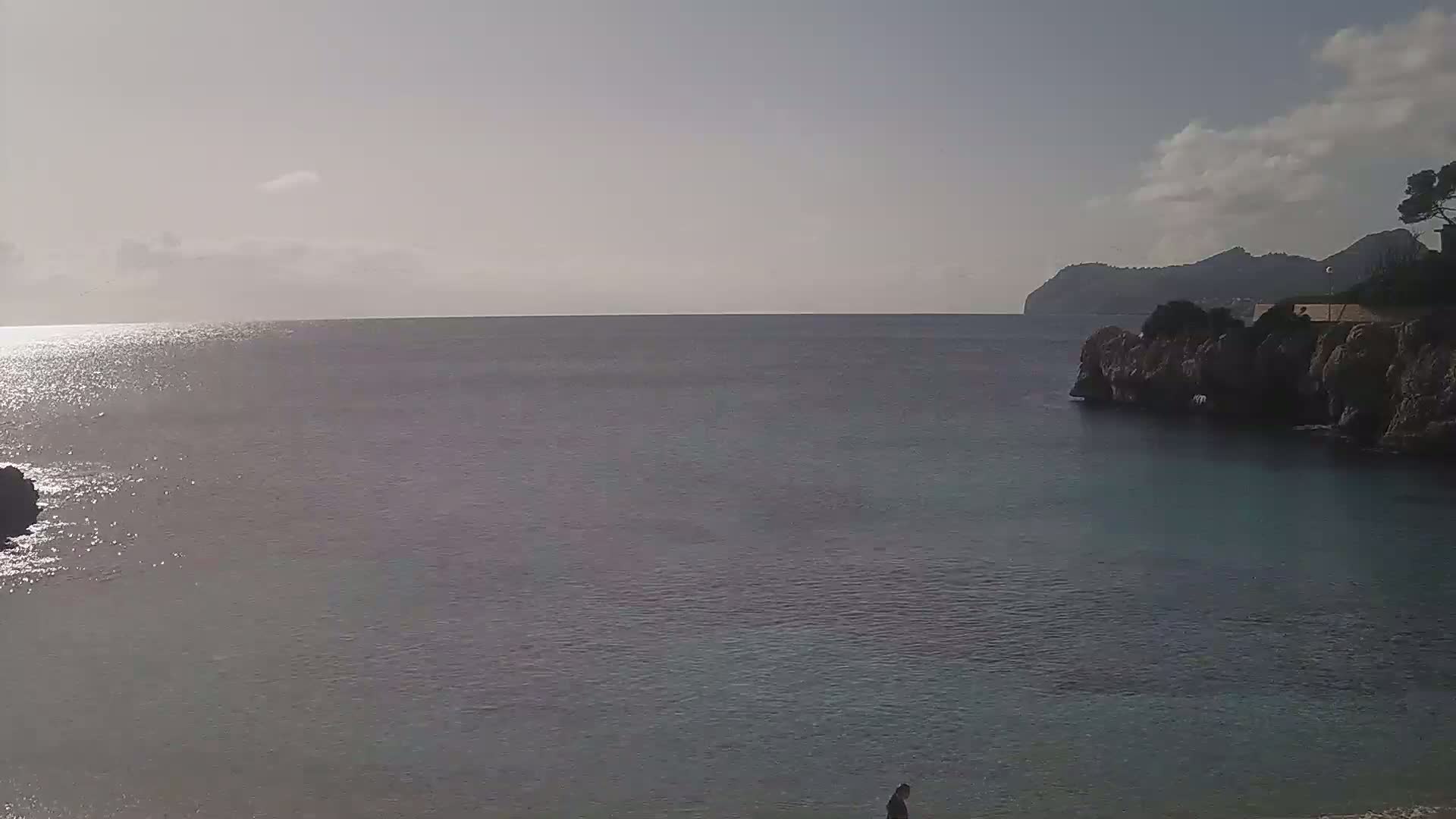 A calm, sunlit sea stretches to a distant mountain range under a partly cloudy sky.