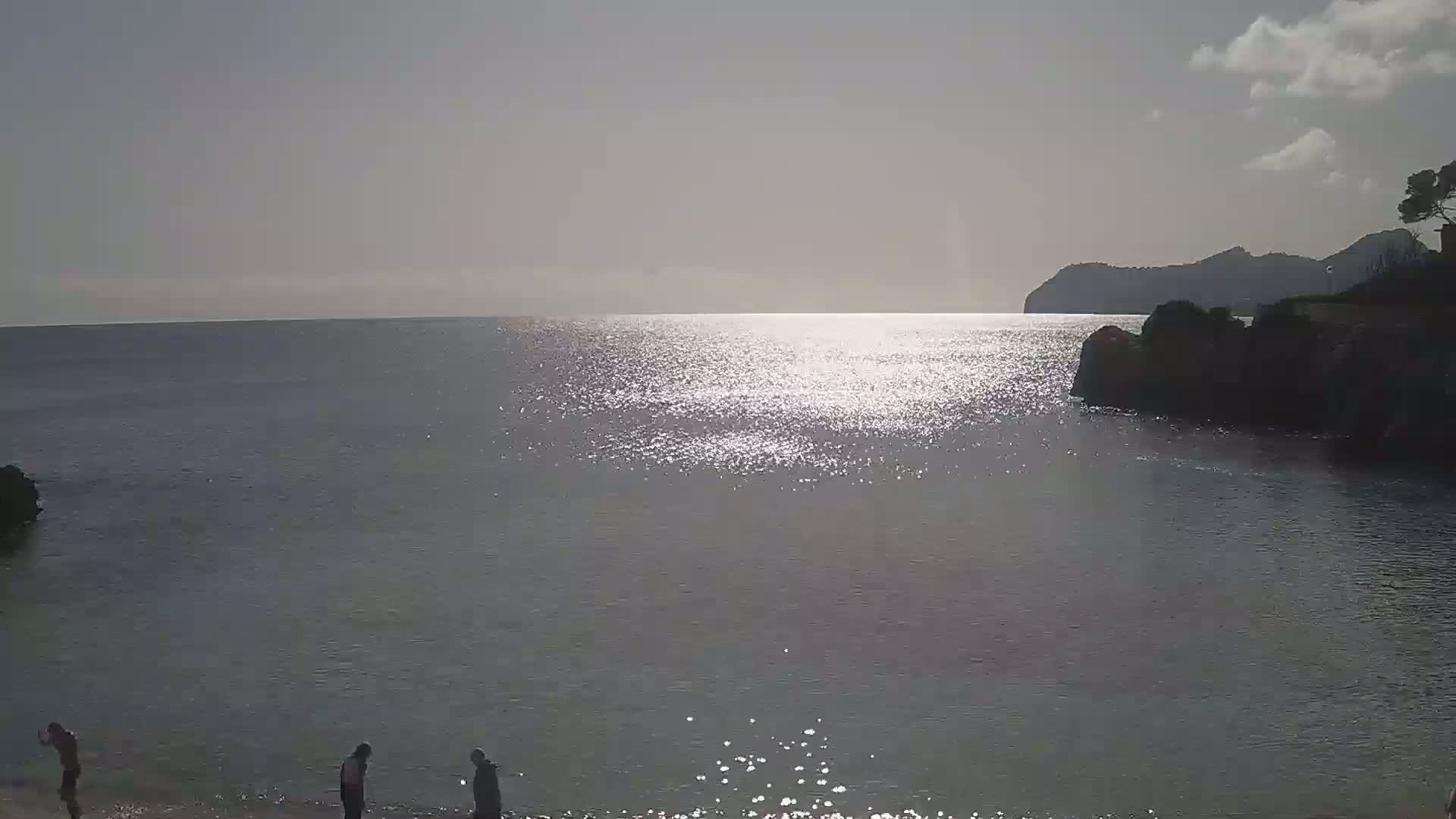 A calm, sunlit sea laps against a rocky shore where a few people stand near the water's edge.