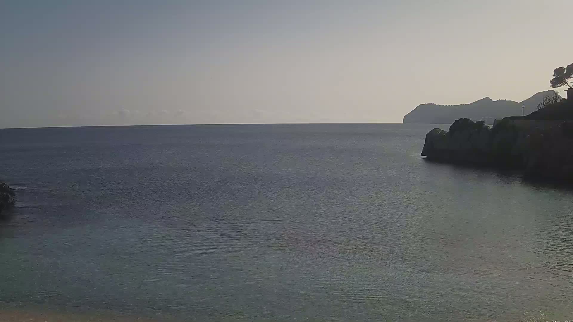 A calm, dark-blue sea stretches to a distant, hazy coastline under a clear, pale sky.
