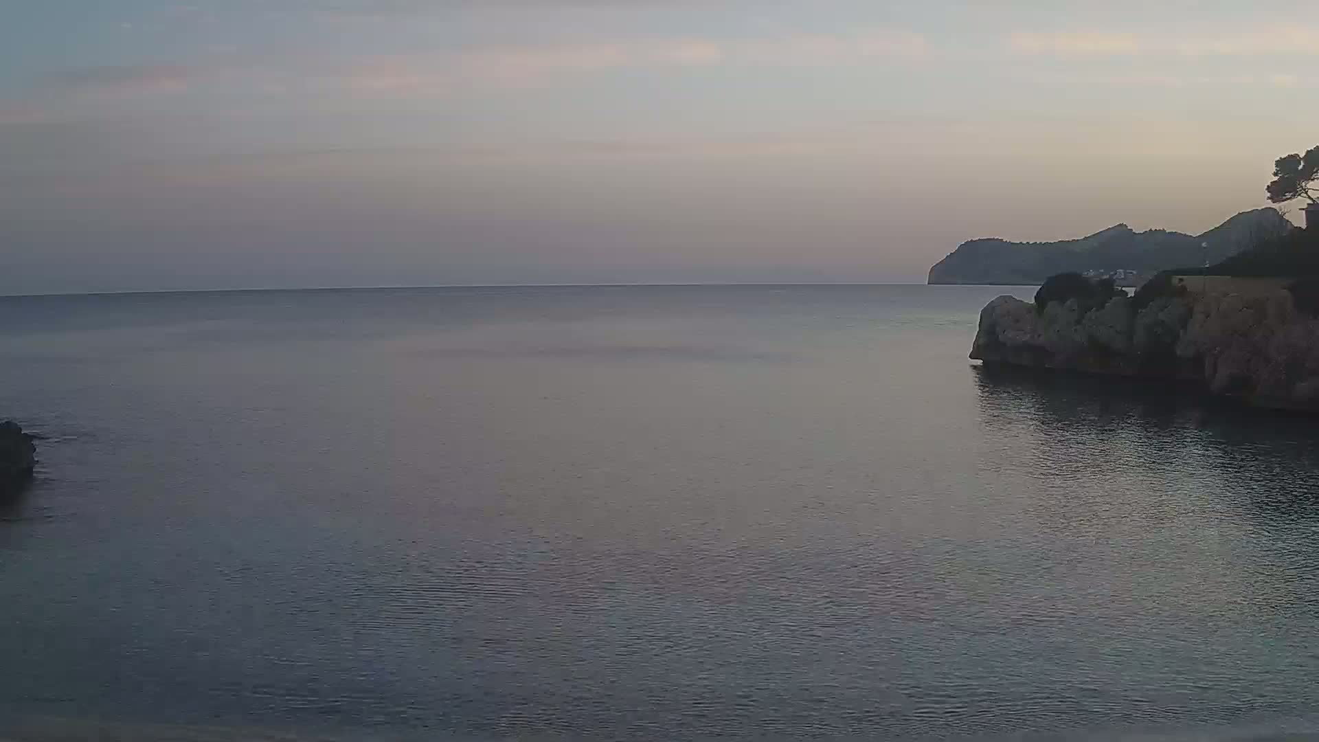 A calm, twilight sea laps against a rocky shoreline, with a distant hill visible under a pale sky.
