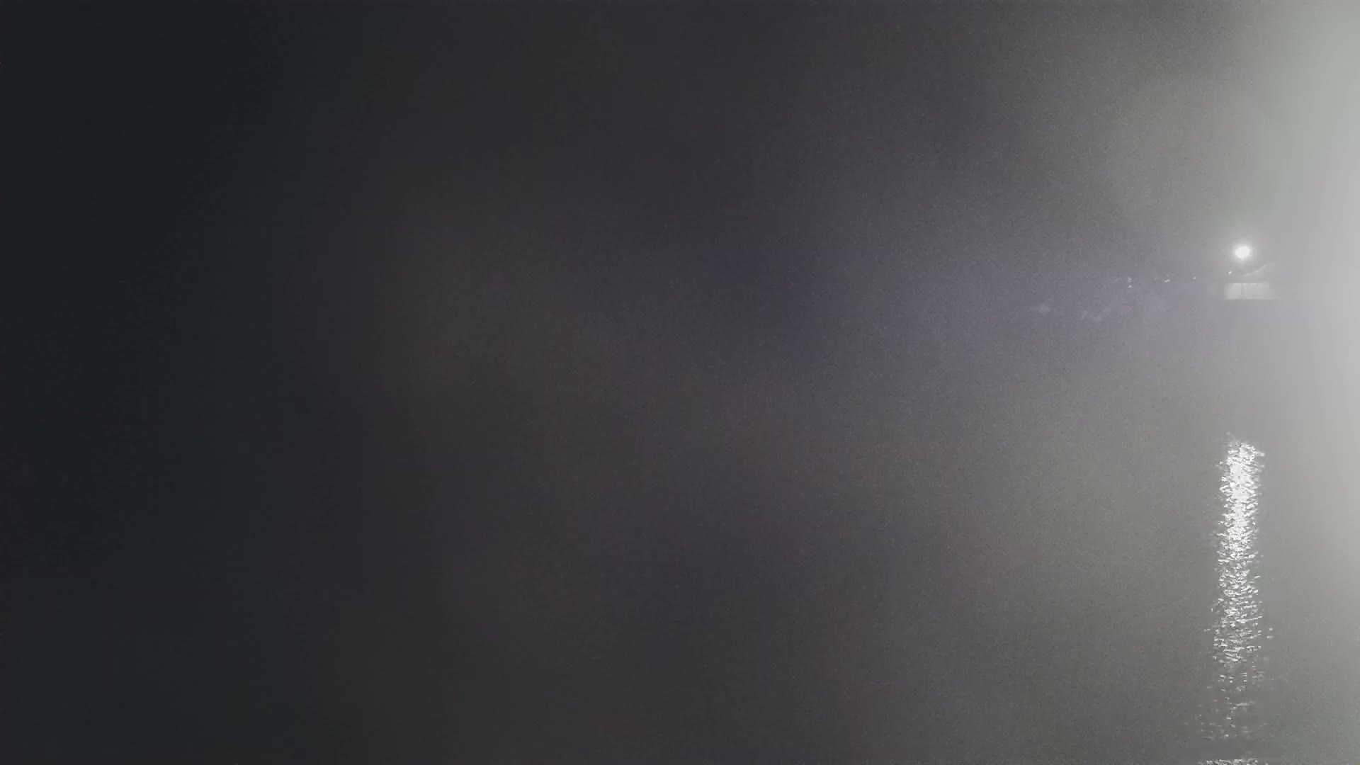 A foggy nighttime scene shows a body of water reflecting a light source in the distance.