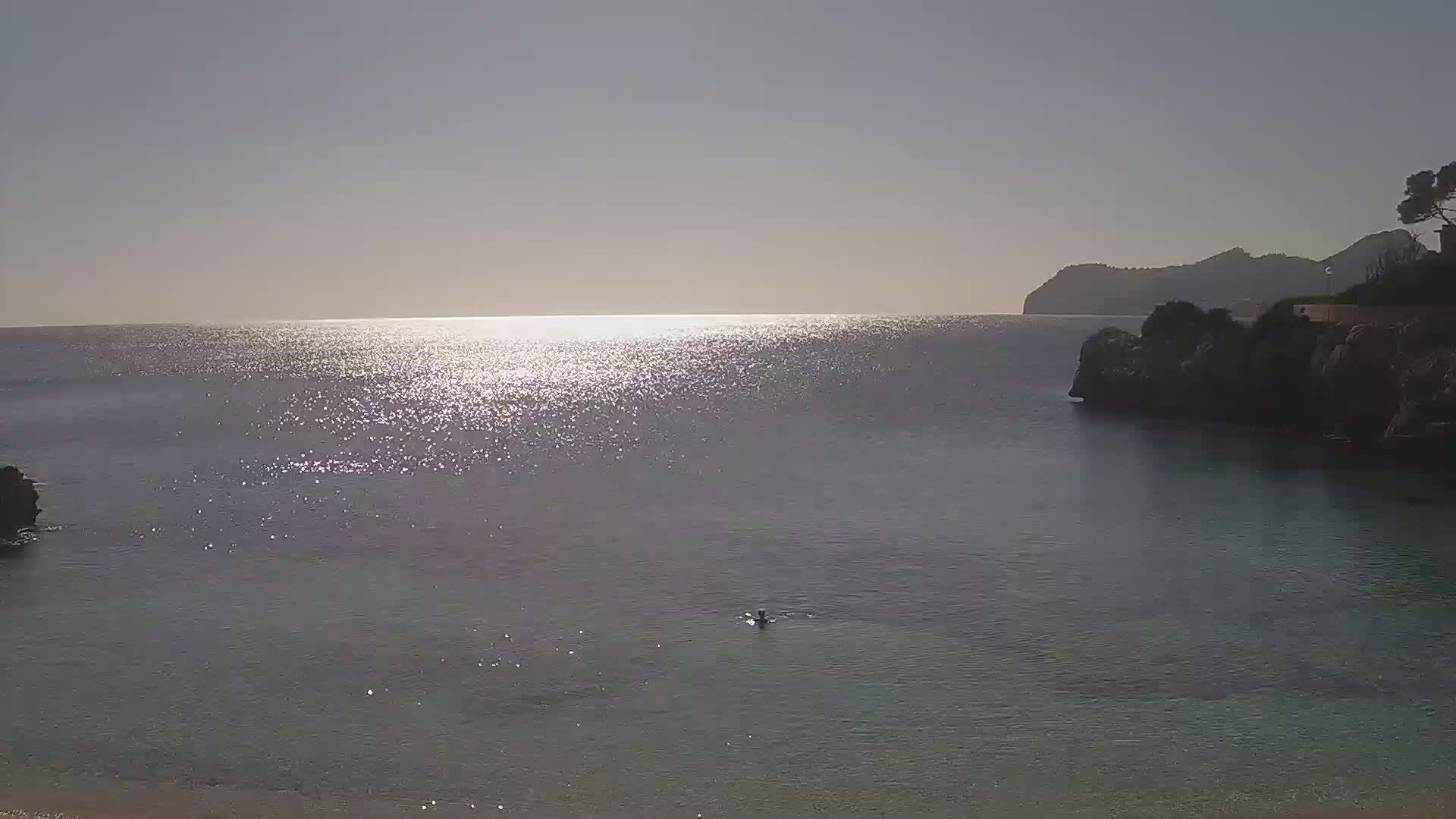 A calm, sunlit sea is partially enclosed by rocky headlands, with a single person swimming in the water.