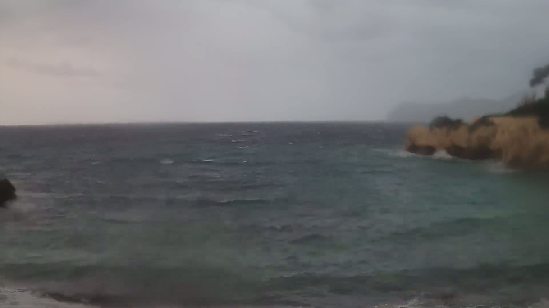 A choppy sea under a gray, overcast sky meets a rocky shoreline with sparse vegetation.