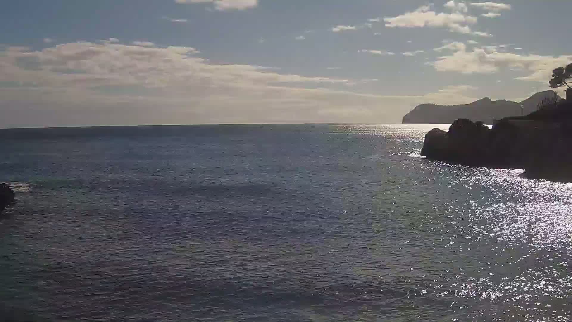 A partly sunny day shows a calm sea reflecting sunlight near a rocky shore with a distant mountain range.