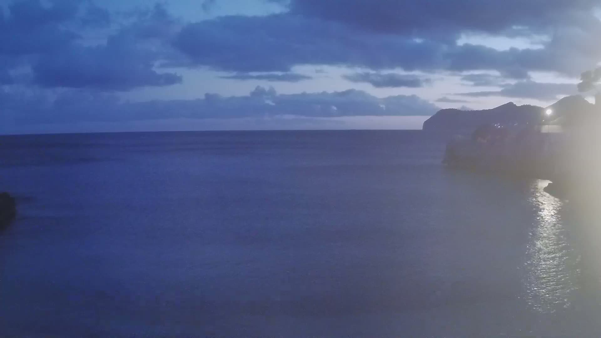 A dark, calm ocean stretches to a distant, silhouetted coastline under a cloudy, twilight sky.