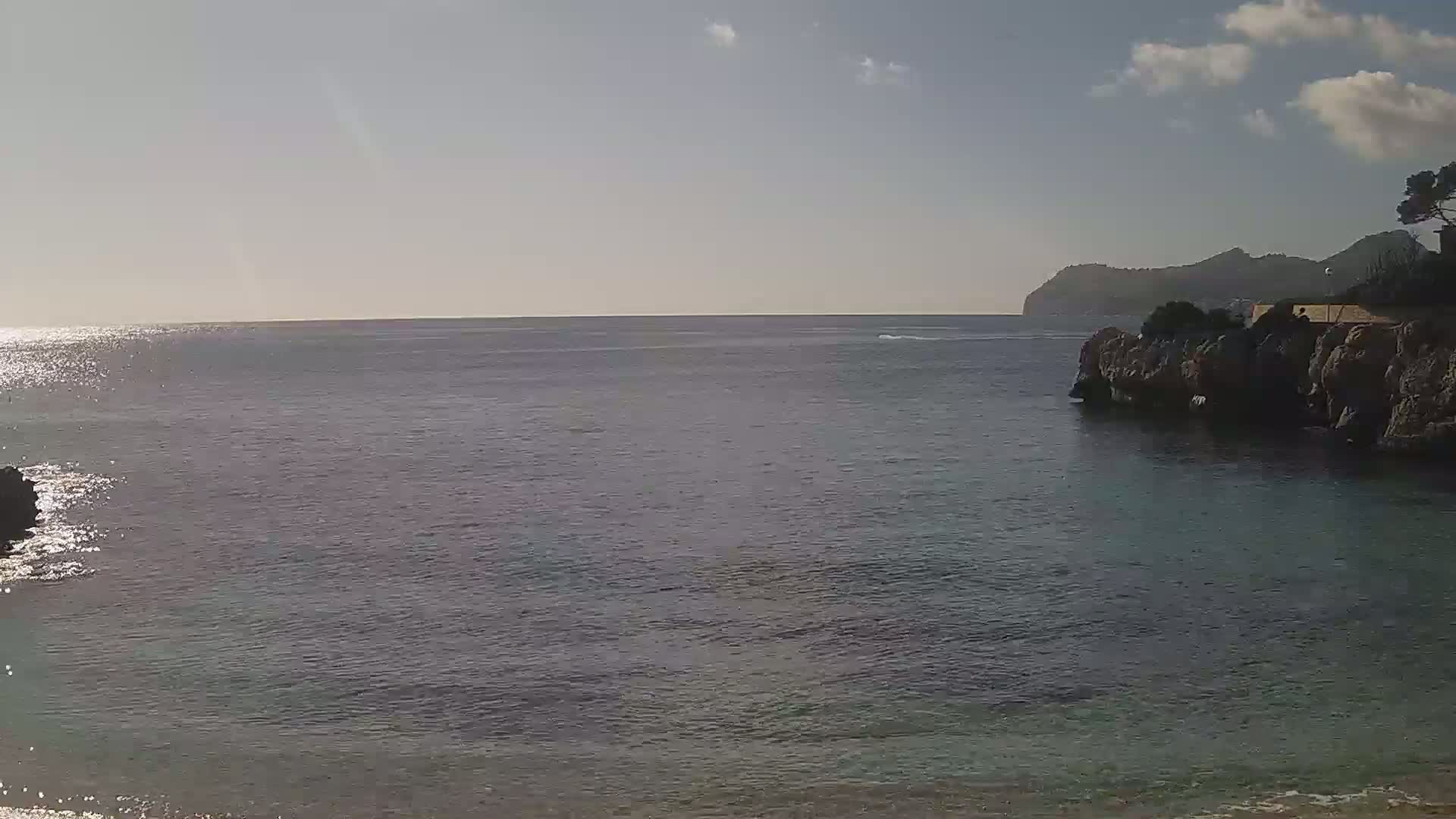 A calm sea meets a rocky shoreline under a partly cloudy sky, with a distant mountain visible.