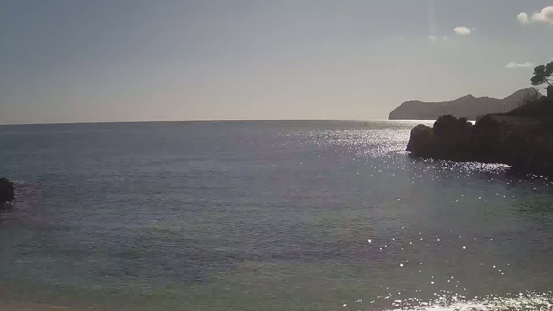 Sparkling water laps against a rocky shore under a mostly clear sky, with a distant landmass visible on the horizon.
