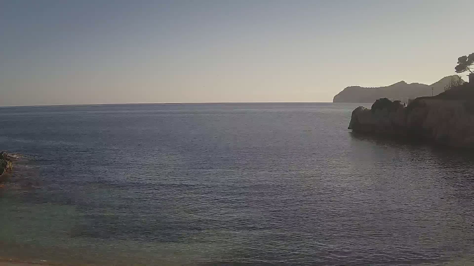A calm, dark-blue sea laps against a rocky shoreline under a clear, pale sky, with a distant mountain visible.