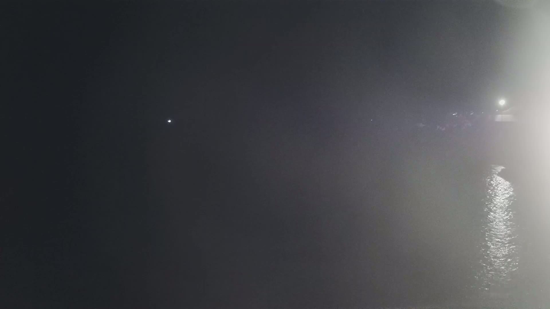 A dark, foggy night scene shows a body of water reflecting light, with a dimly lit shoreline in the distance.