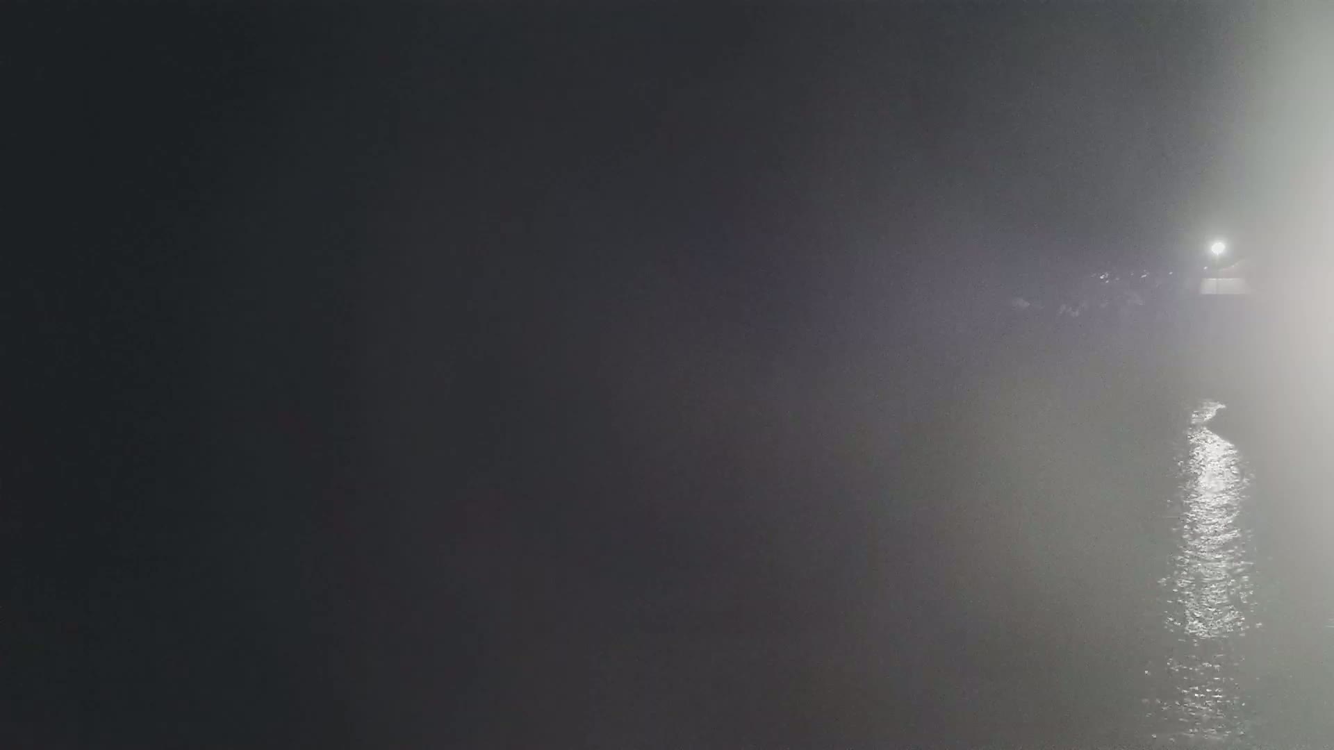 A dimly lit body of water at night is partially obscured by fog or mist, with a light source visible in the distance.