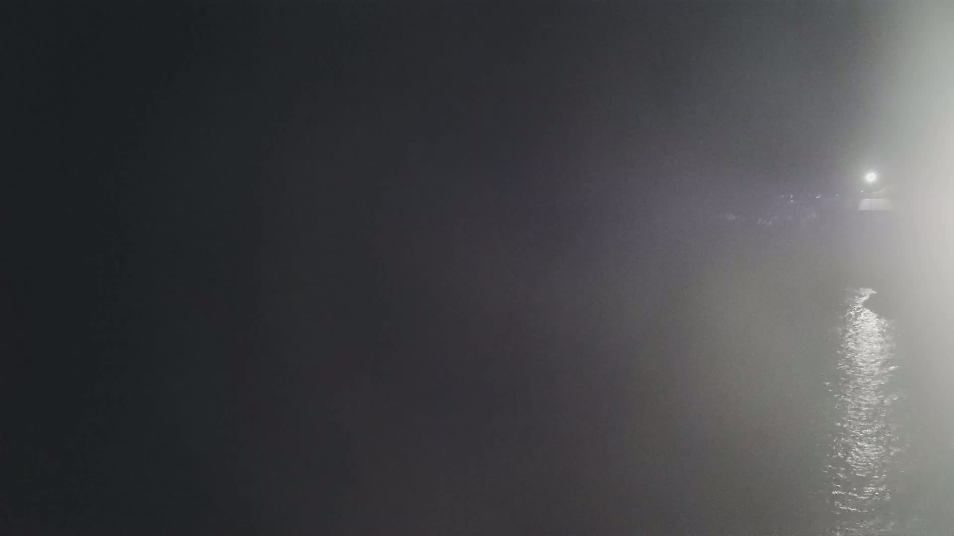 A dimly lit body of water is seen at night in foggy conditions, with a light source visible in the distance.
