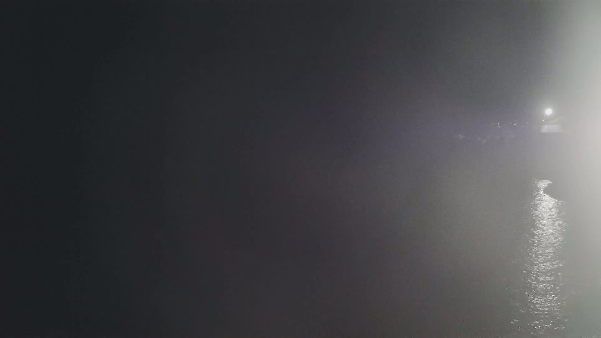 A dimly lit body of water is shrouded in thick fog at night, with a faint light visible in the distance.