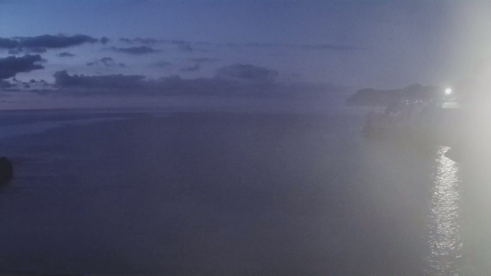A hazy, dark-blue coastal scene at twilight shows a body of water with a distant shoreline partially obscured by fog.