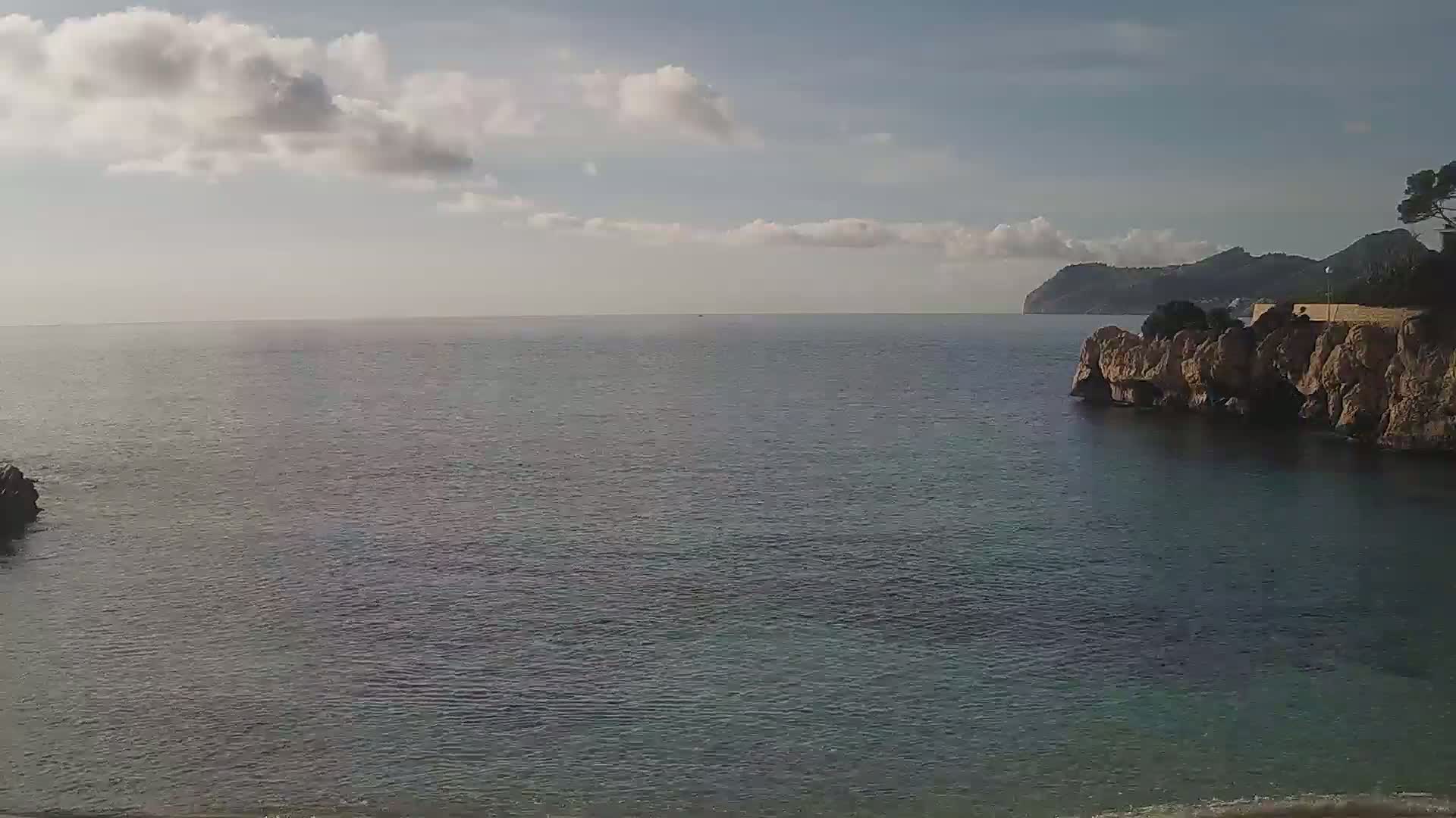 A calm, partly cloudy day shows a view of a rocky coastline with a dark blue sea extending to a distant mountain range.