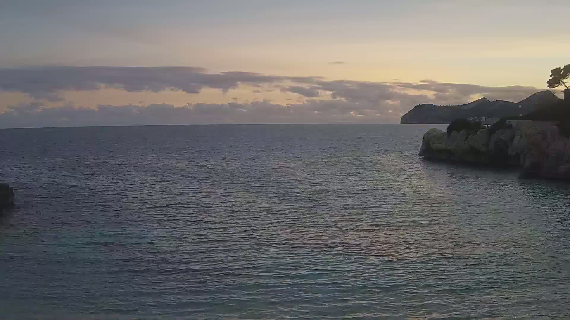 A calm sea at dawn, under a mostly cloudy sky, is seen from a rocky cove with a distant mountain in the background.