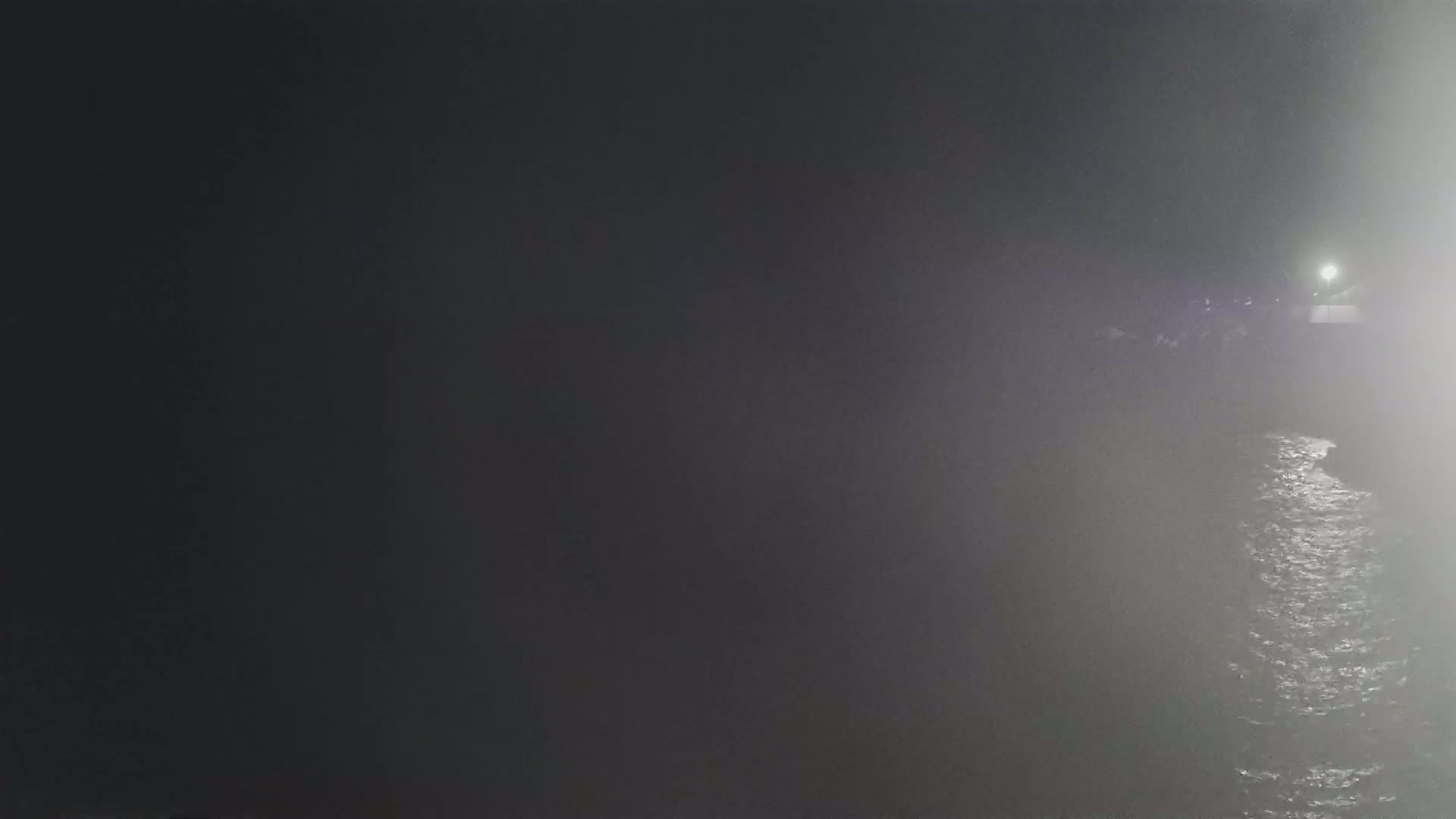 A foggy nighttime scene shows dark land meeting a body of rippling water, illuminated by a single light source in the distance.