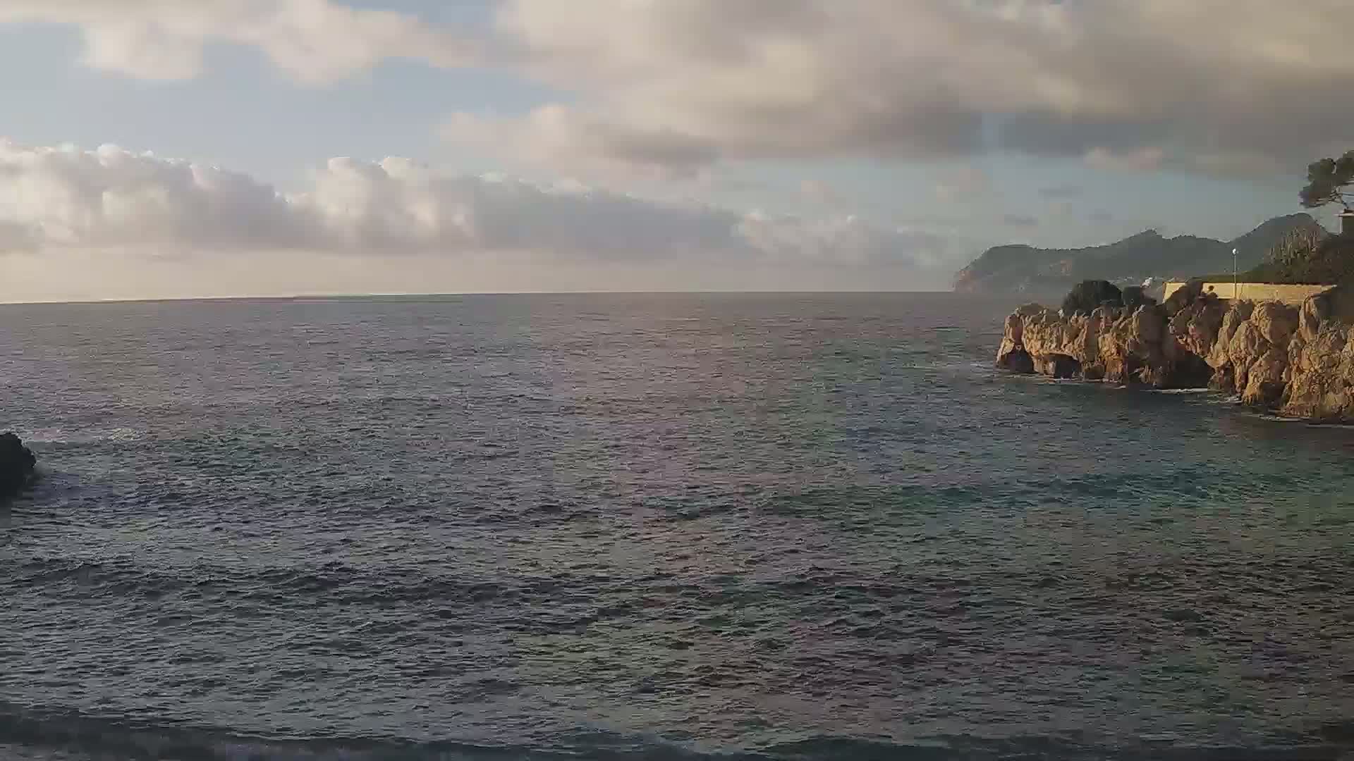 A partly cloudy sky overlooks a calm sea meeting a rocky, cliff-side shore.