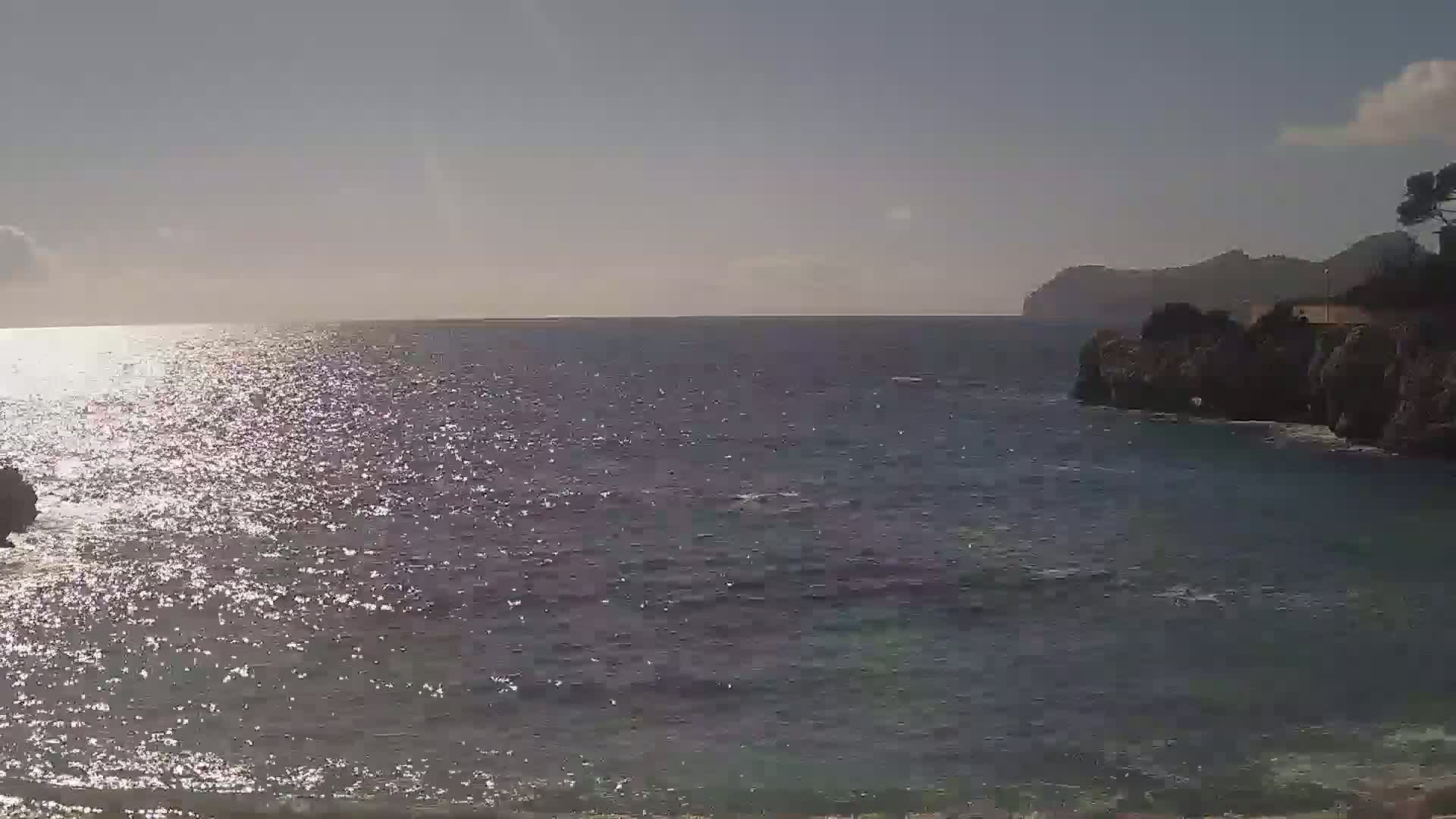 A sparkling sea under a mostly clear sky stretches to a distant mountain range, with rocky cliffs and sparse vegetation visible along the shoreline.